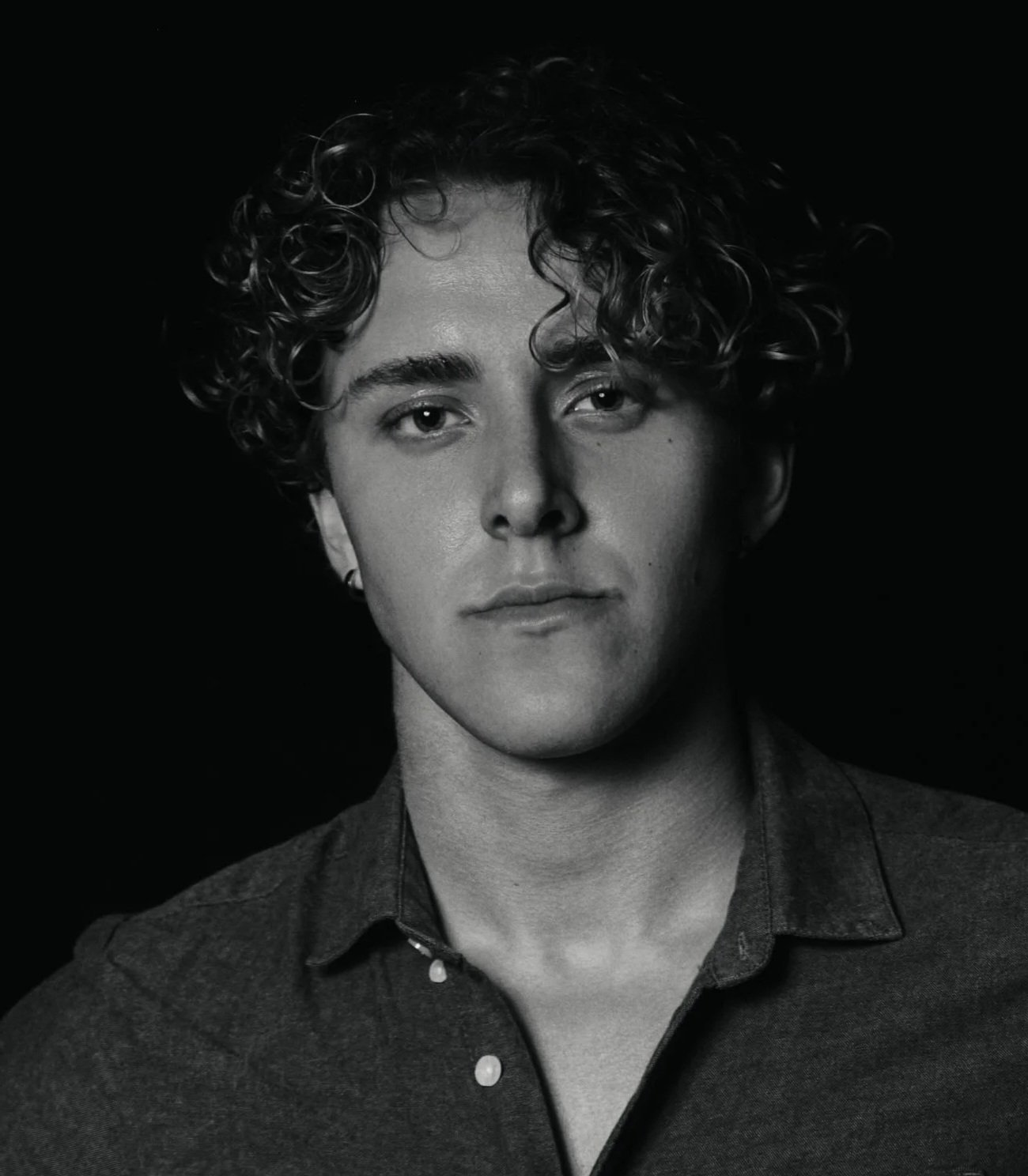 Black and white portrait of a young man with curly hair, looking directly at the camera, wearing a button-up shirt.