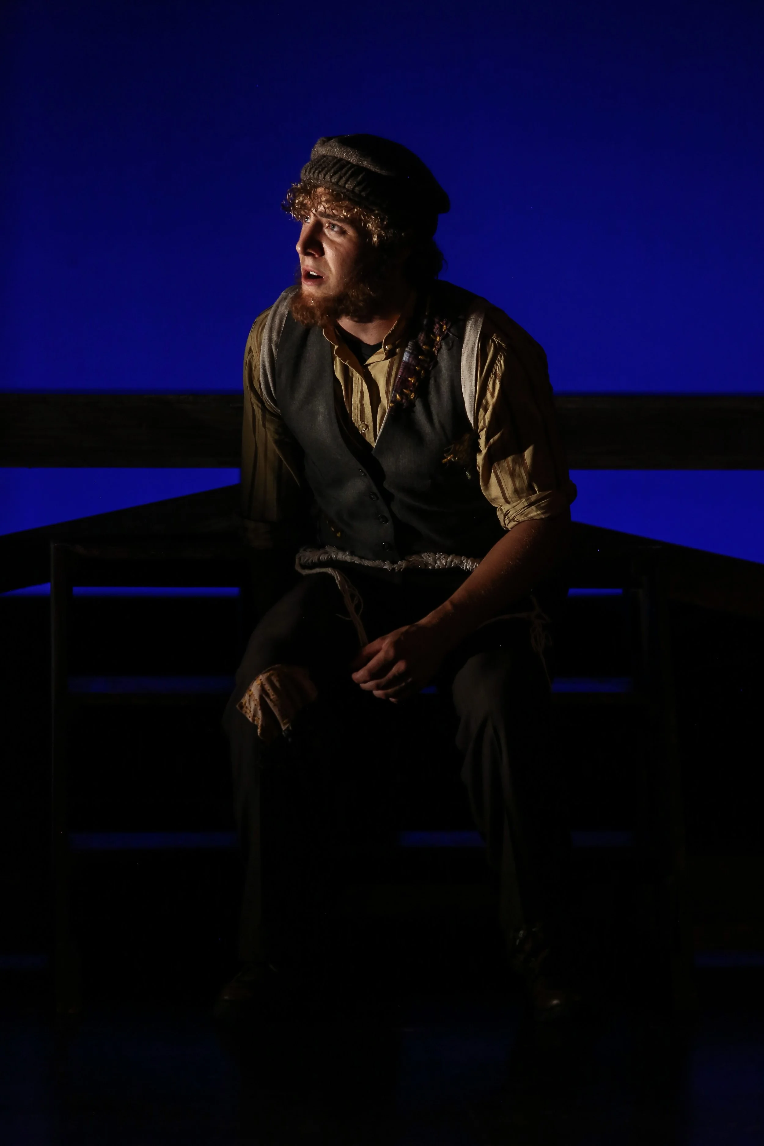 A young man with curly hair and a beard, wearing a flat cap and vintage clothing, sitting on a wooden bench against a dark blue background, appearing to be in deep thought or performing in a theatrical scene.