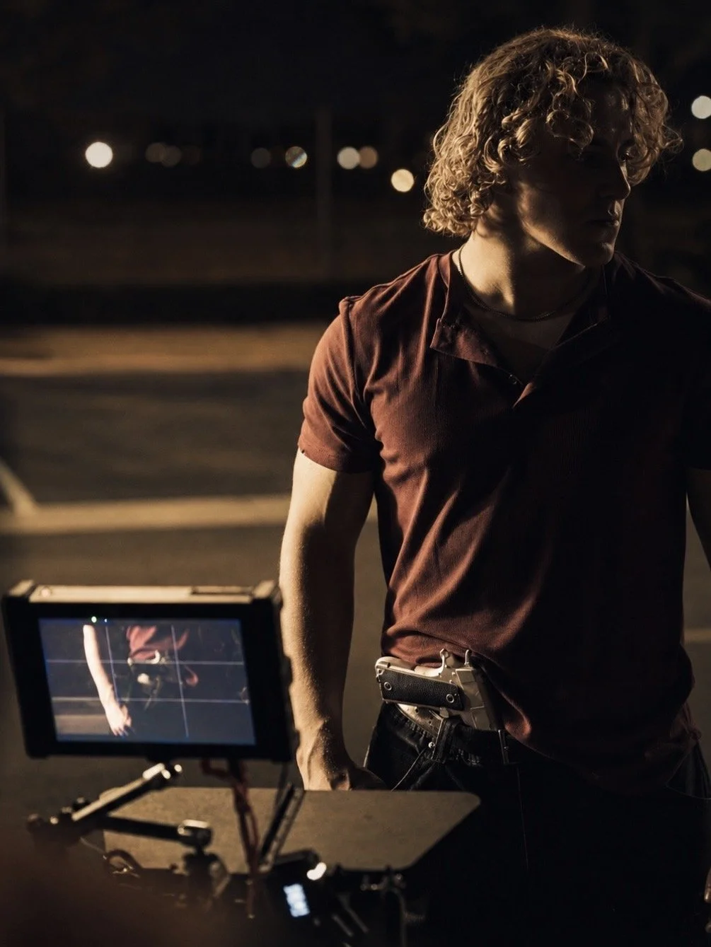 A young man with curly blond hair standing in a dimly lit outdoor area at night, with a monitor in front of him displaying his reflection.