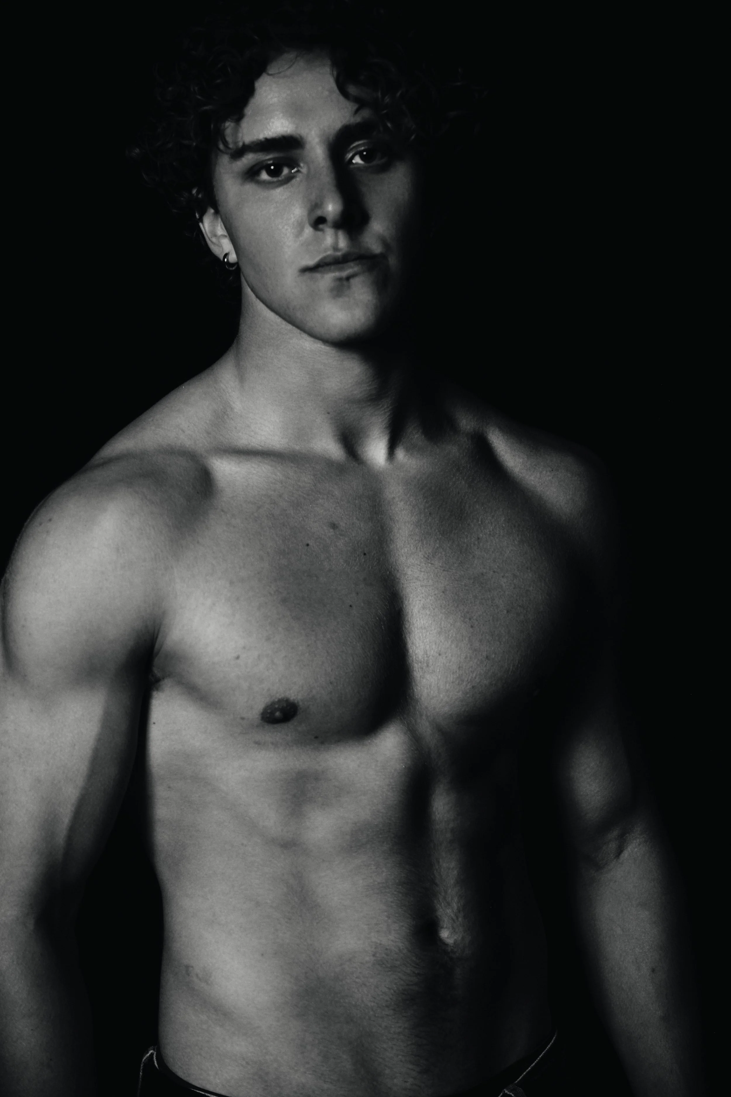A black and white photo of a topless person with curly hair, wearing earrings and a bracelet, with a serious expression, against a solid dark background.