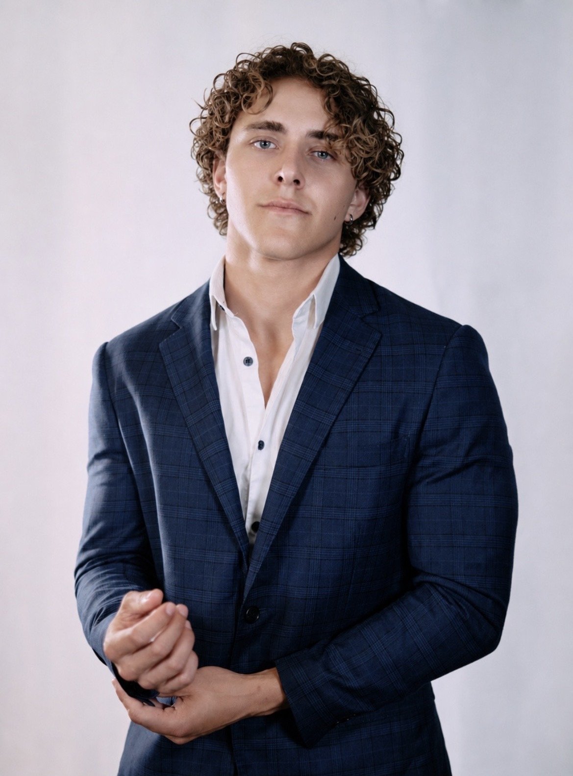 A young man with curly hair, light skin, and blue eyes wearing a dark blue checkered suit jacket and a white shirt, standing against a plain background.