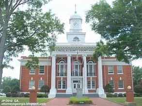 Historic brick courthouse with white columns, clock tower, and steps, surrounded by trees.