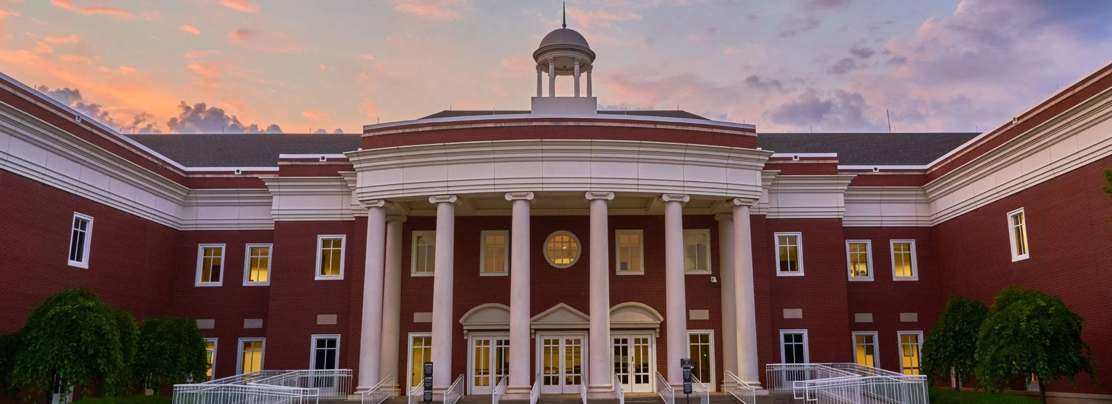 A large brick building with white columns and a dome on top, during sunset with a colorful sky.