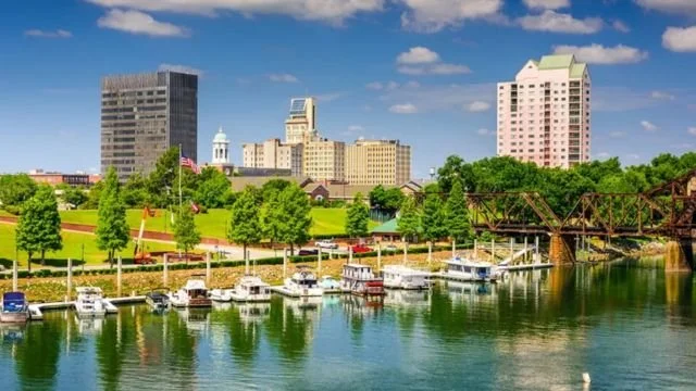 City skyline with tall buildings and a park in front, overlooking a marina with boats on a river under a partly cloudy blue sky.