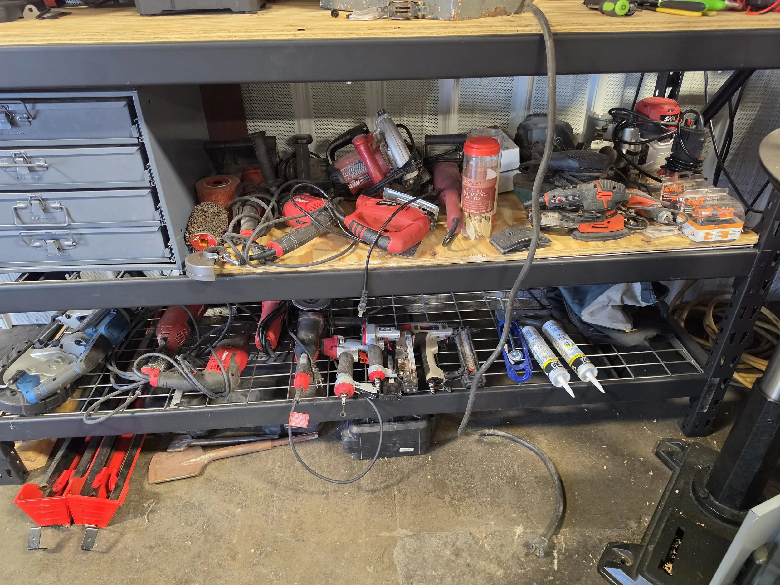 A workbench with various power tools, hand tools, and supplies, including drills, a grinder, screwdrivers, and a container of biscuits, with a pegboard wall in the background.