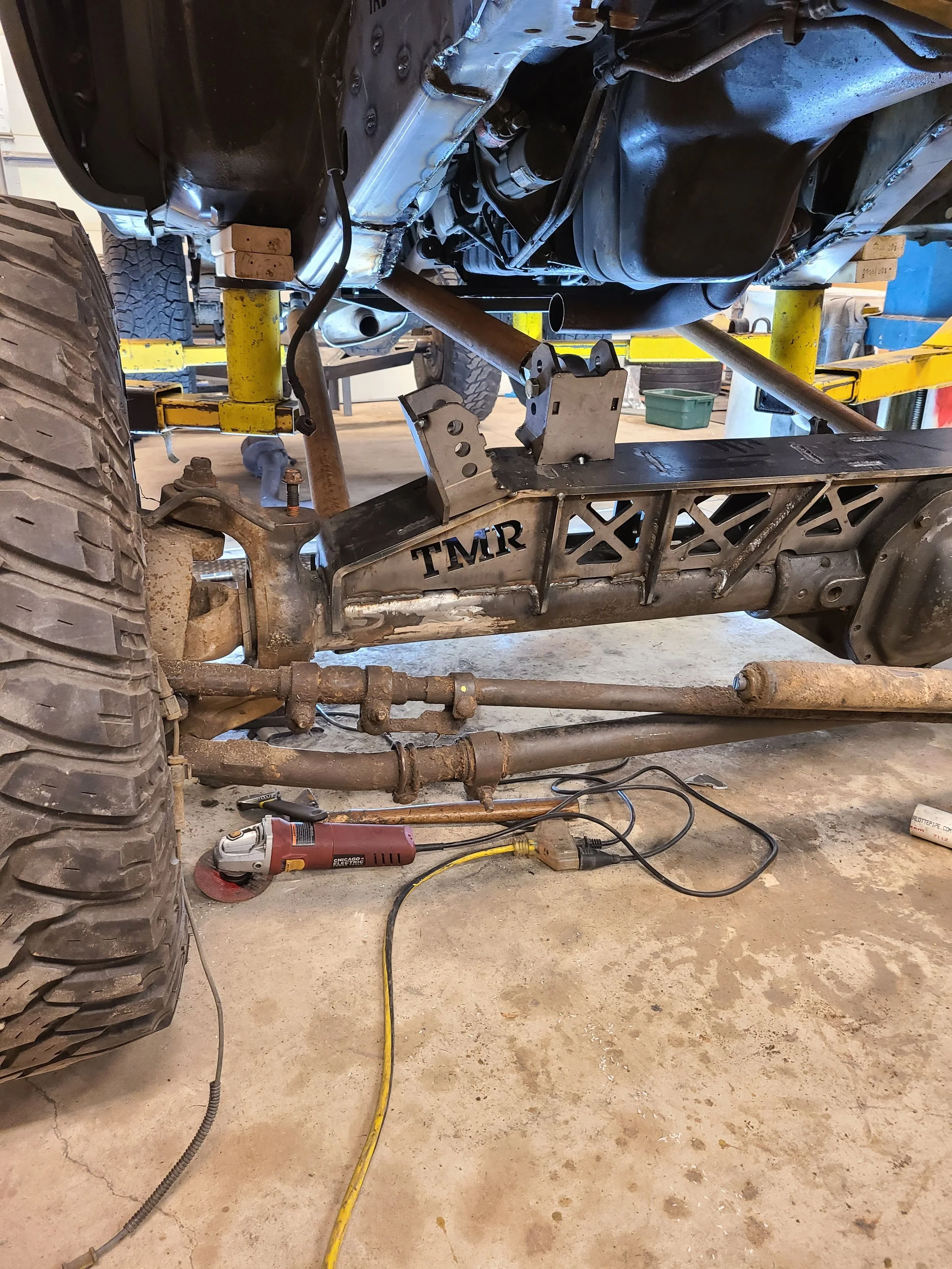 Underneath view of a vehicle in a garage, showing parts of the suspension, chassis, wheels, and tools scattered on the floor, including a rotary tool, a yellow extension cord, and some metal brackets.
