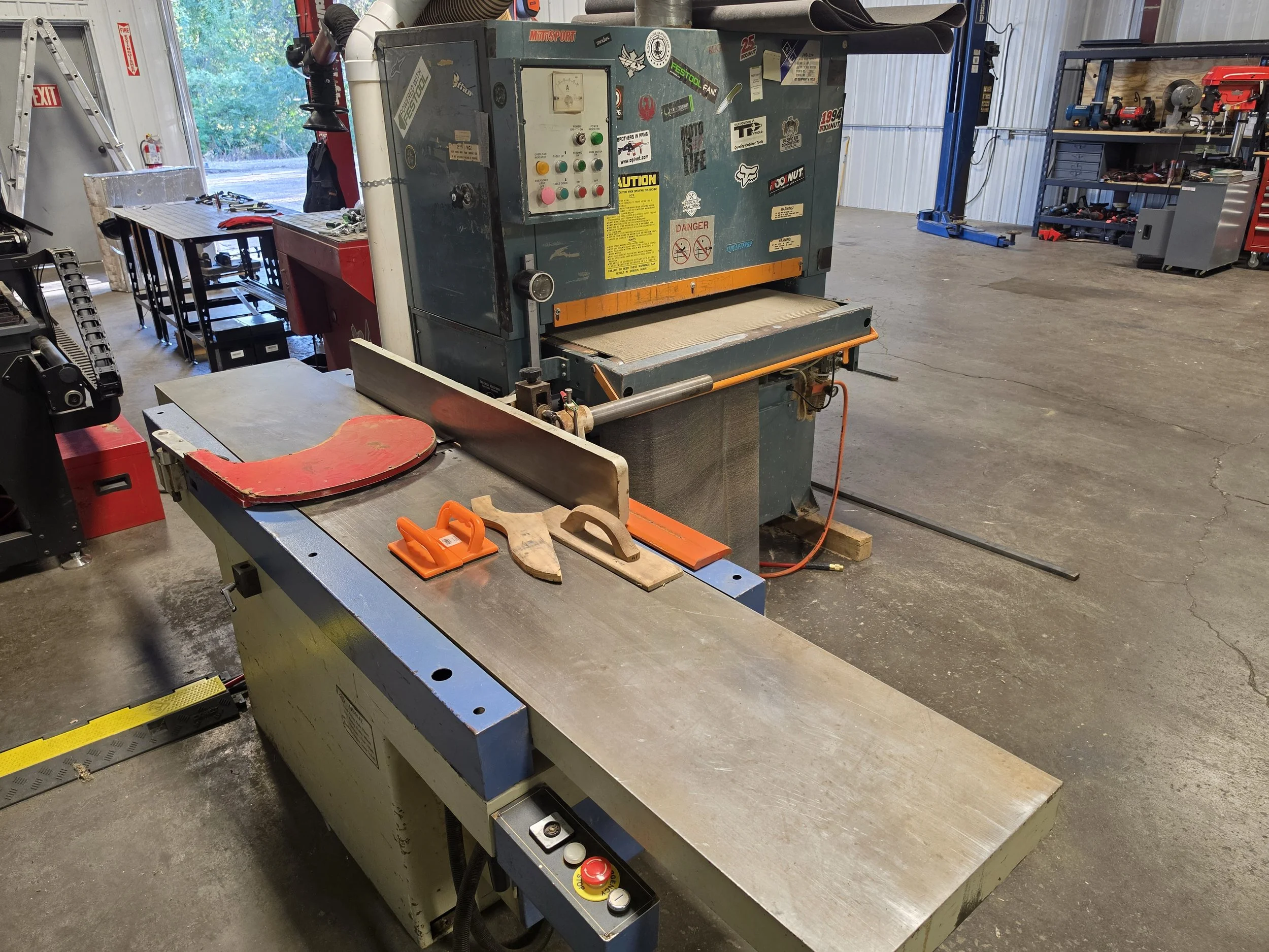 A large industrial machine in a workshop, with various control buttons and labels, surrounded by workbenches, tools, and equipment.