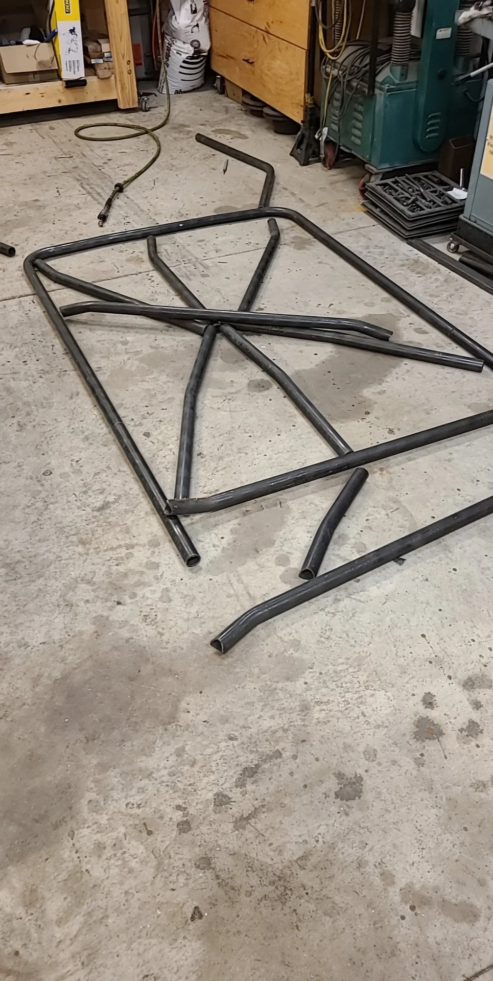 Metal frame parts on concrete floor in a workshop, with tools and equipment in the background.