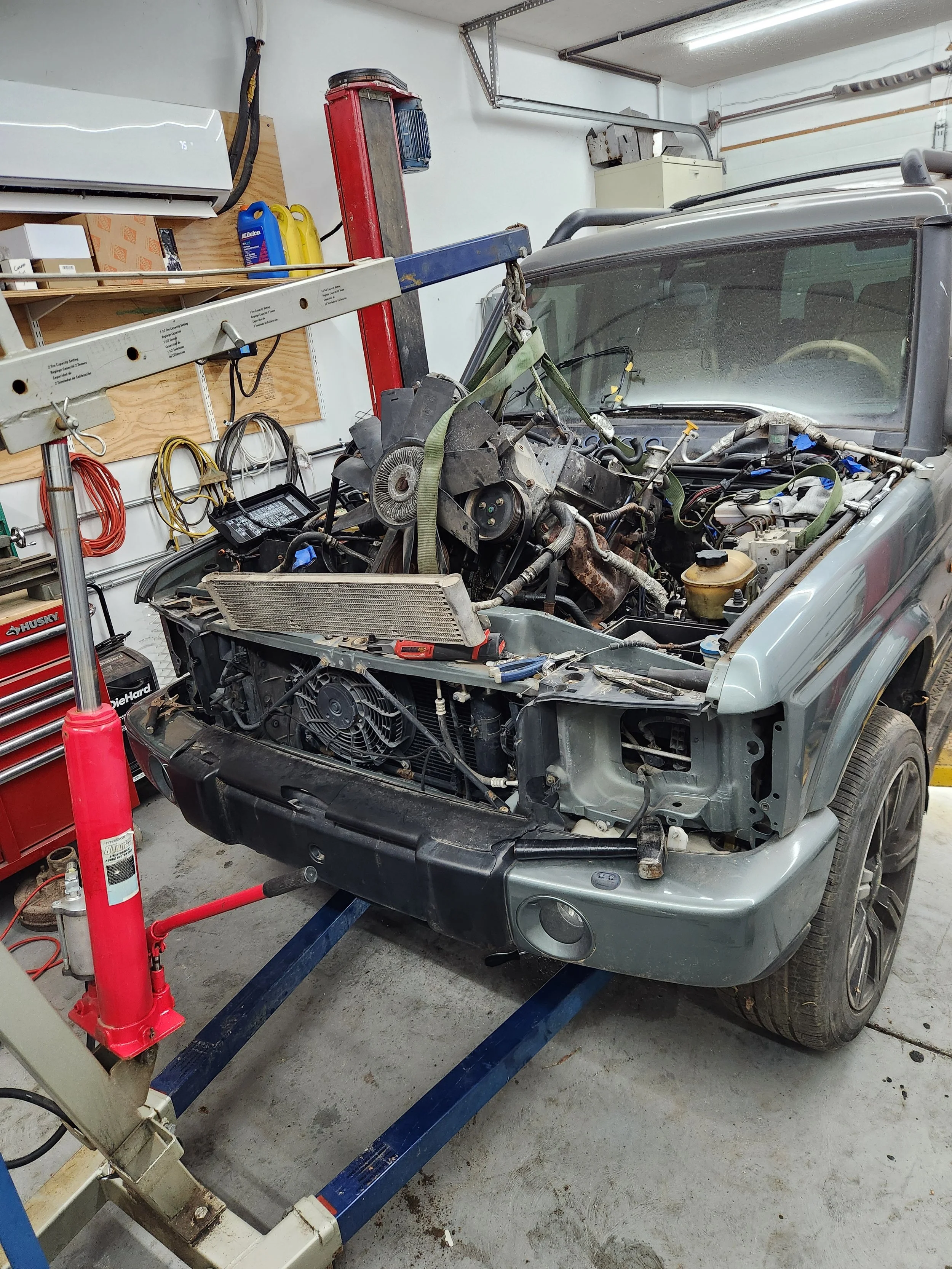 A car with its hood open on a lift in a garage, undergoing engine repairs, with tools and equipment around.