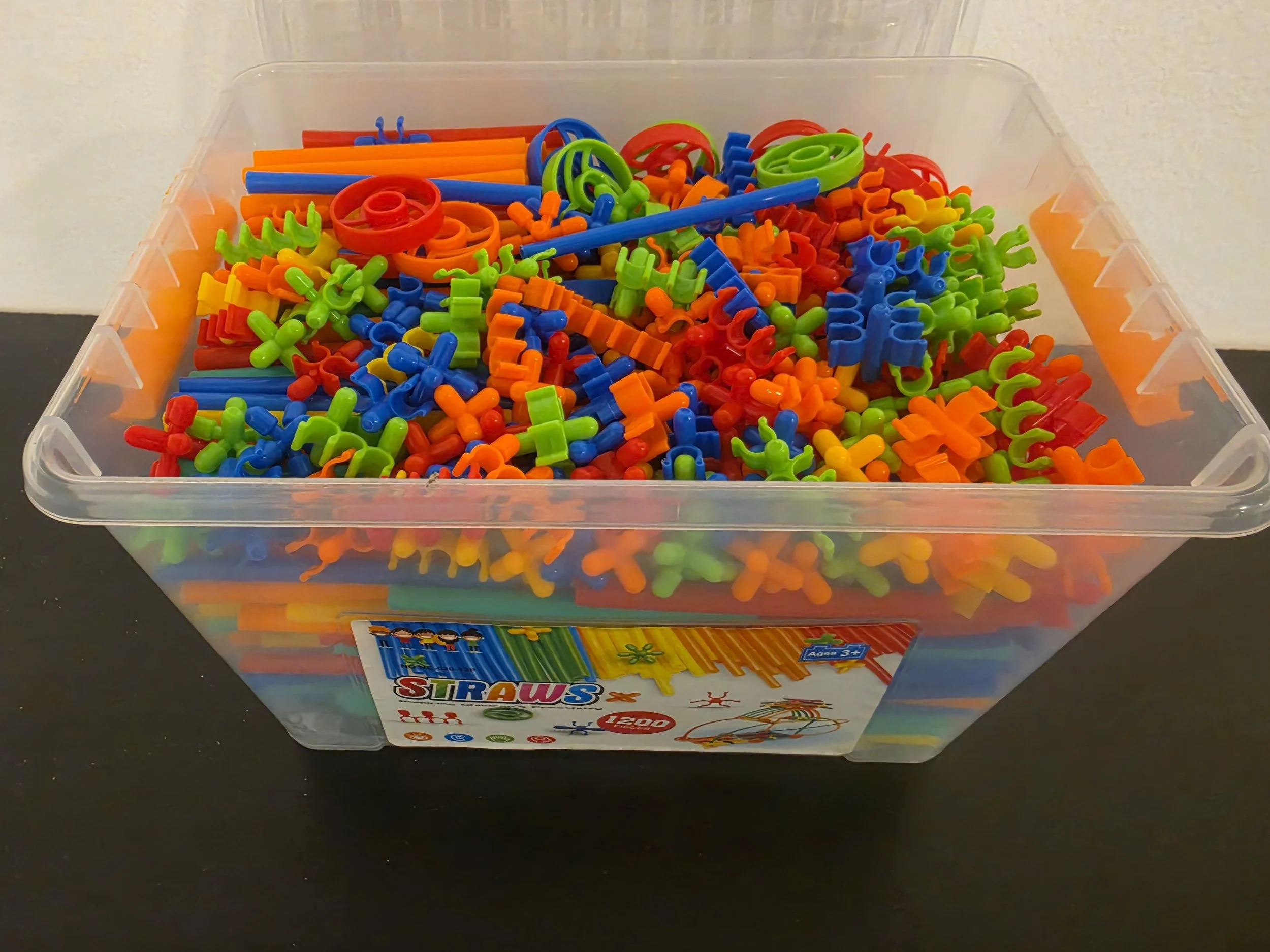 A clear plastic bin filled with colorful rubber straws in blue, green, orange, and red, with a partially visible colorful label on the box.