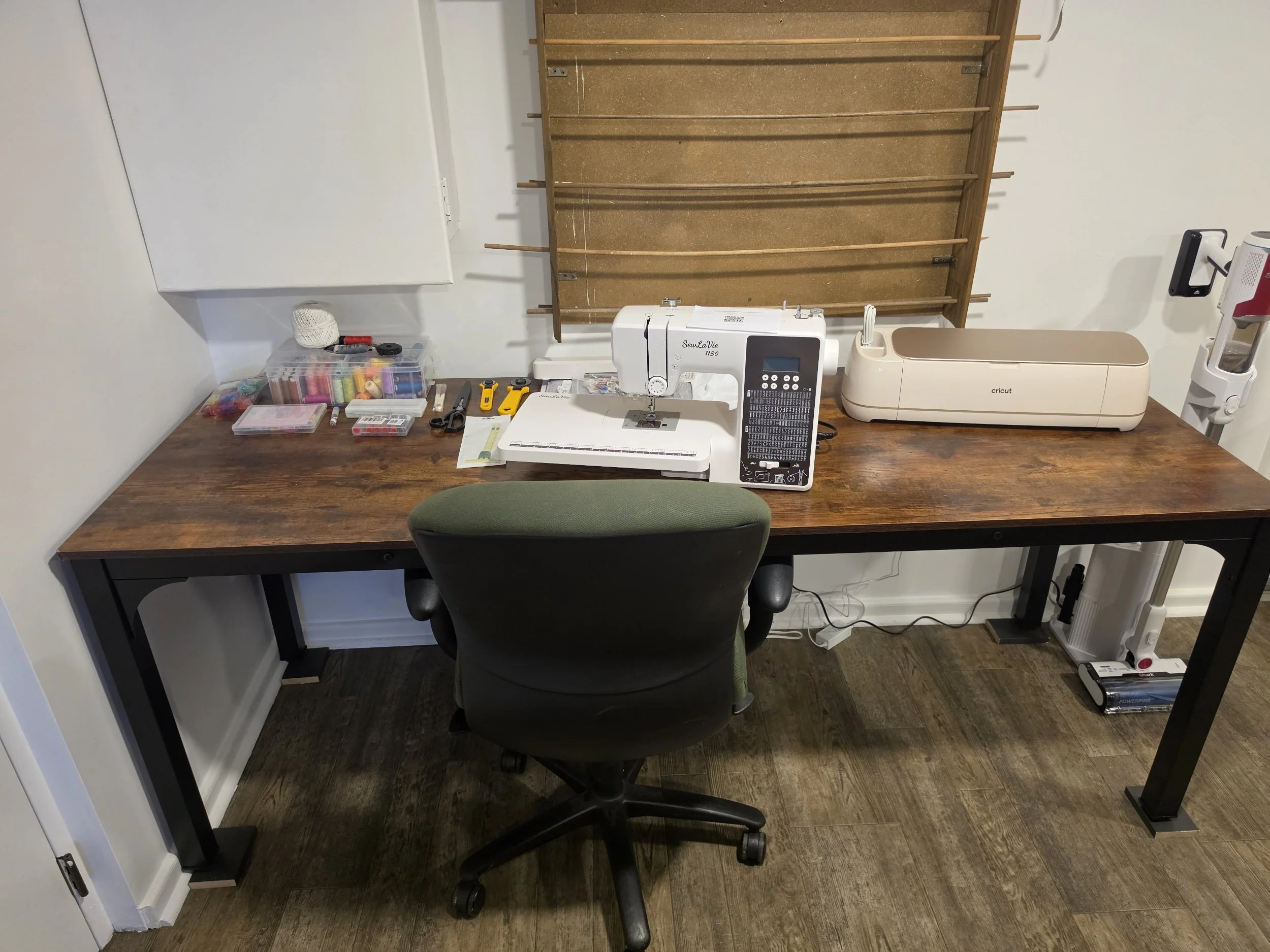 A work desk with a sewing machine, scissors, bobbins, and a plastic container with sewing supplies, a Cricut machine, and a chair in front of the desk, against white walls and a wooden floor.