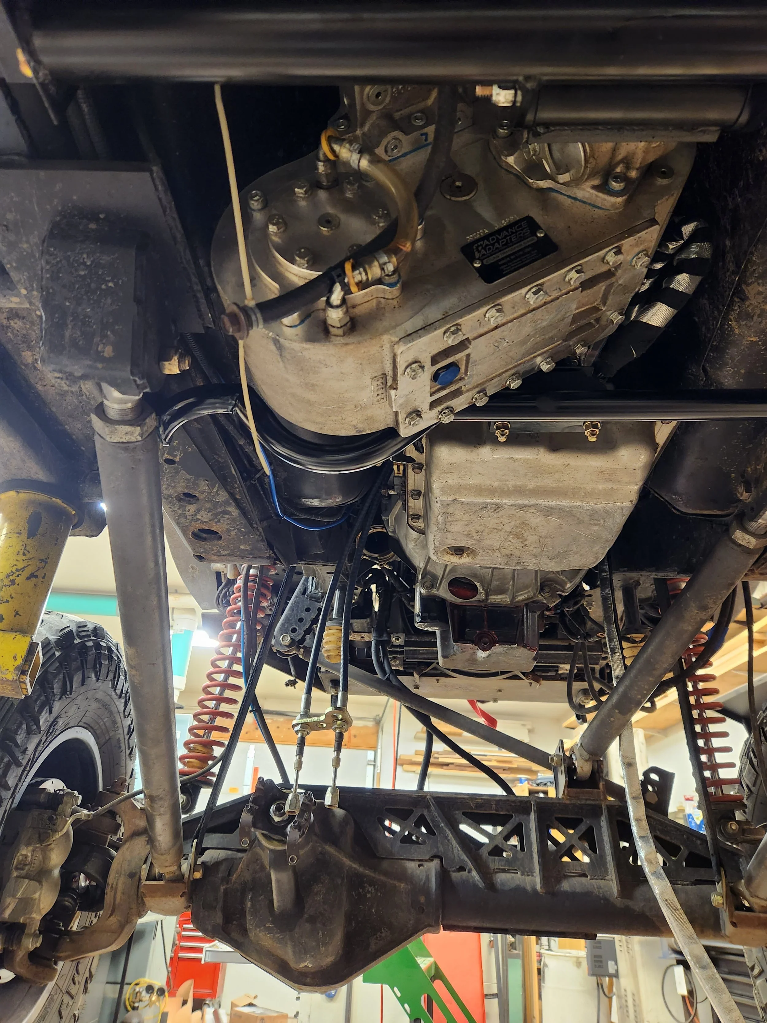 Underneath view of a vehicle's engine compartment showing various mechanical parts, pipes, and wiring, including an oil pan, suspension coils, and engine components in a workshop setting.