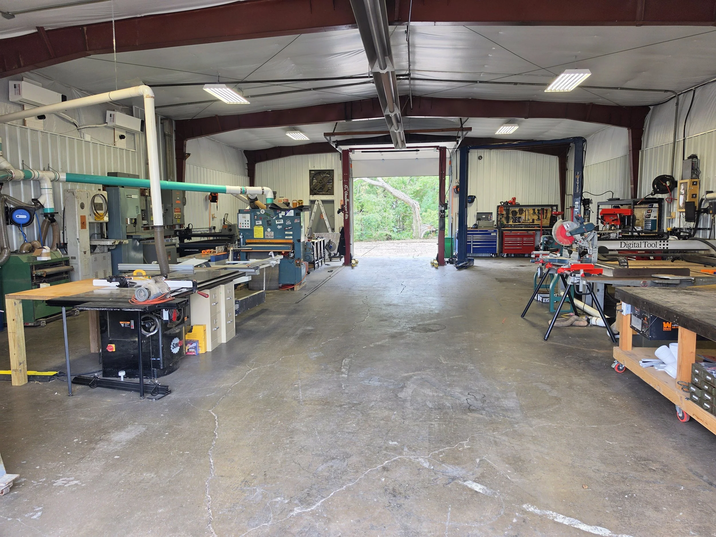 Interior view of a workshop or garage with various tools and equipment, including saws, workbenches, and storage units, with an open door revealing a green outdoor area.