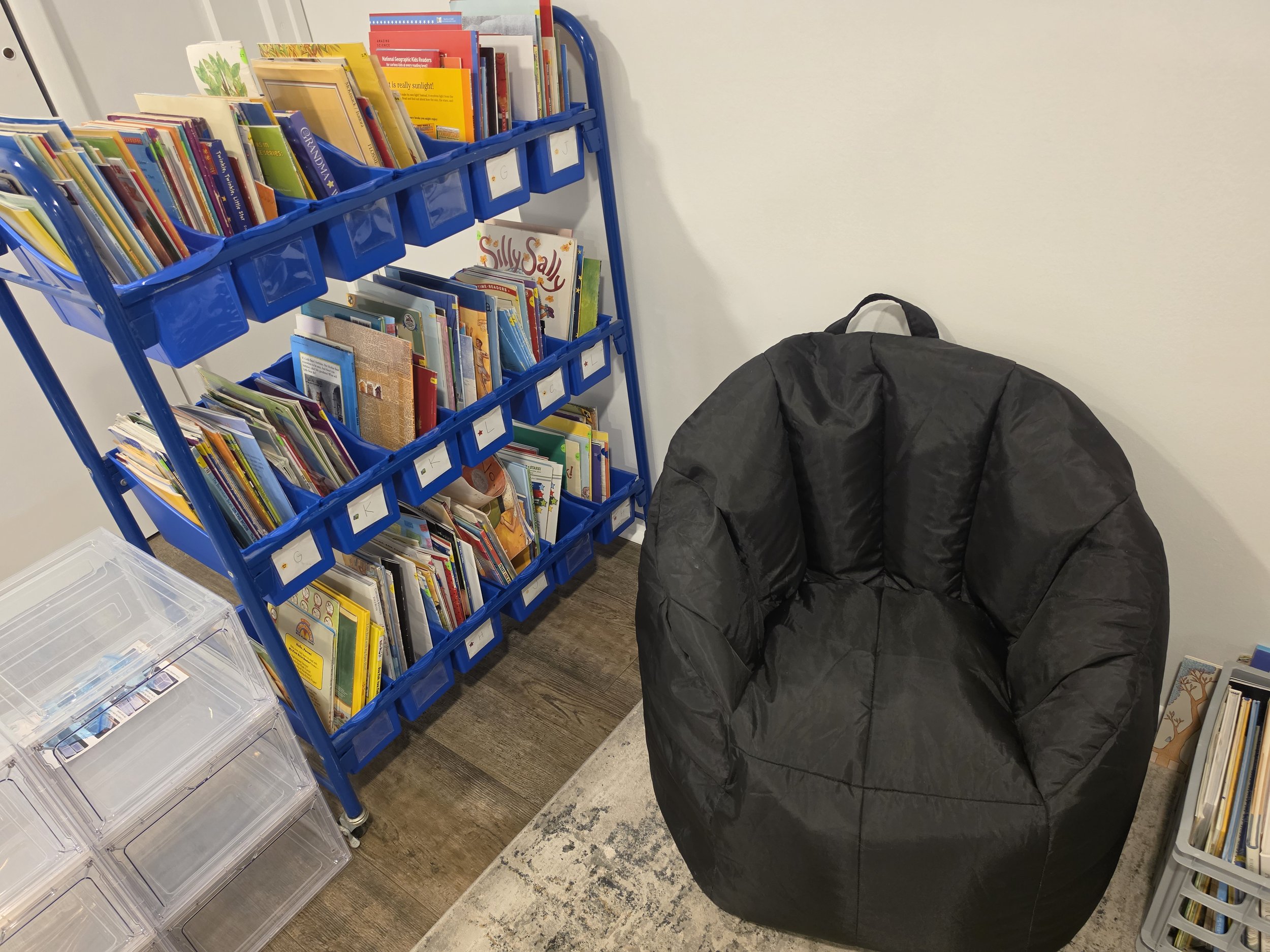 A blue bookshelf filled with children's books, next to a black bag and clear plastic storage boxes in a room with wooden flooring.