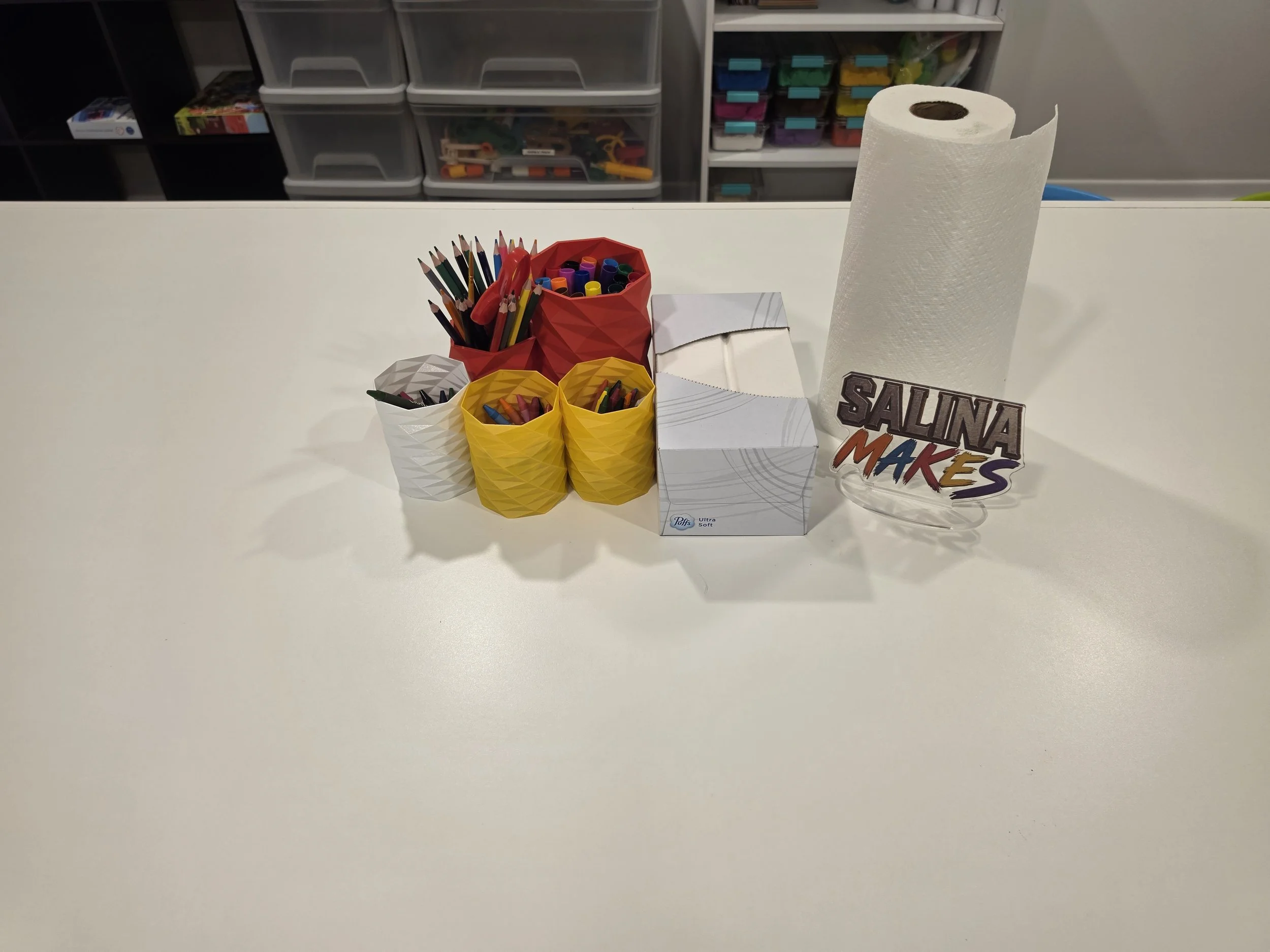 A white table with colorful craft supplies, including colored pencils, markers, and paper towels, with storage bins filled with various arts and crafts items in the background.