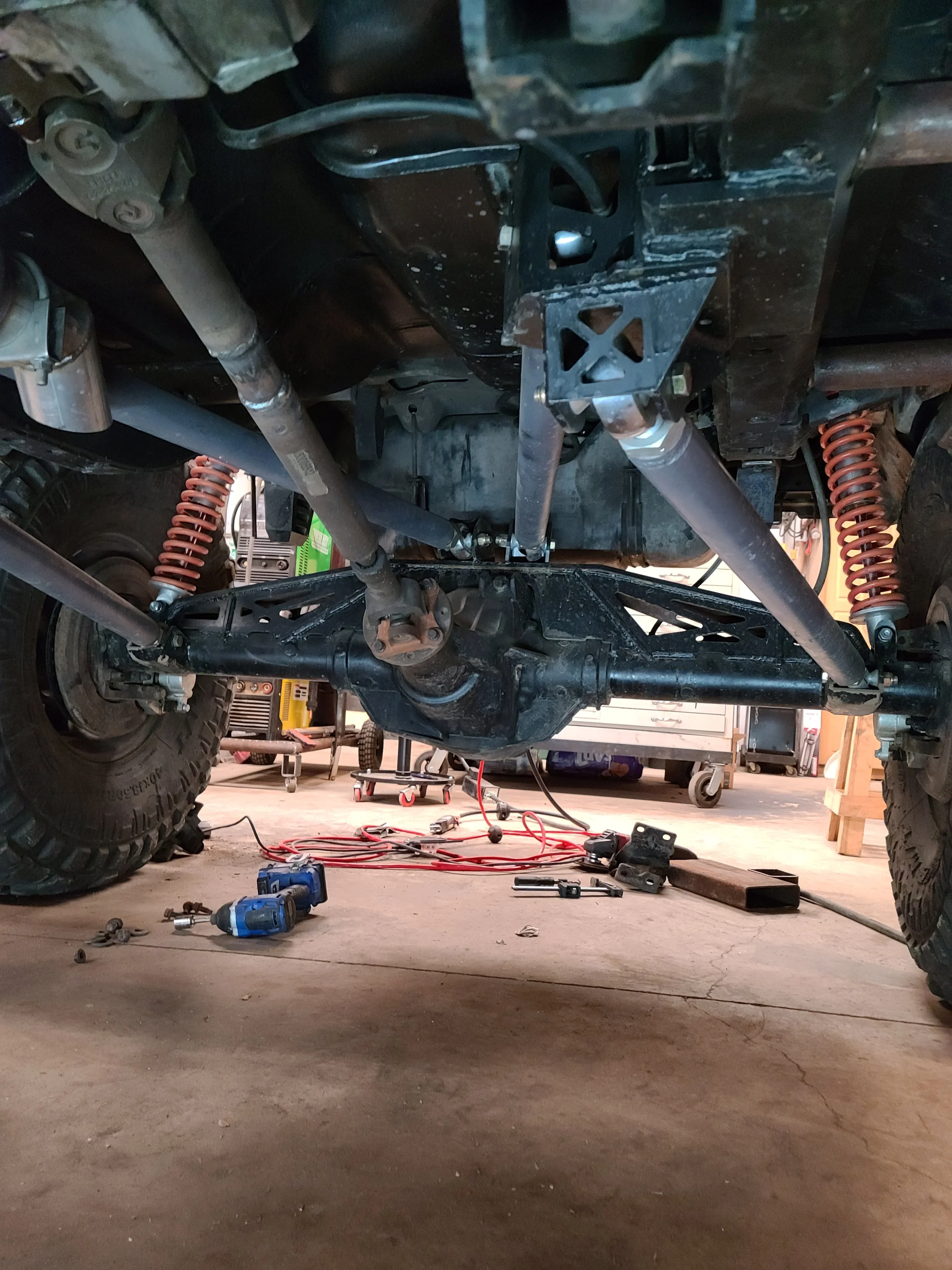 Underneath view of a vehicle's suspension and chassis with tools and parts on the garage floor.
