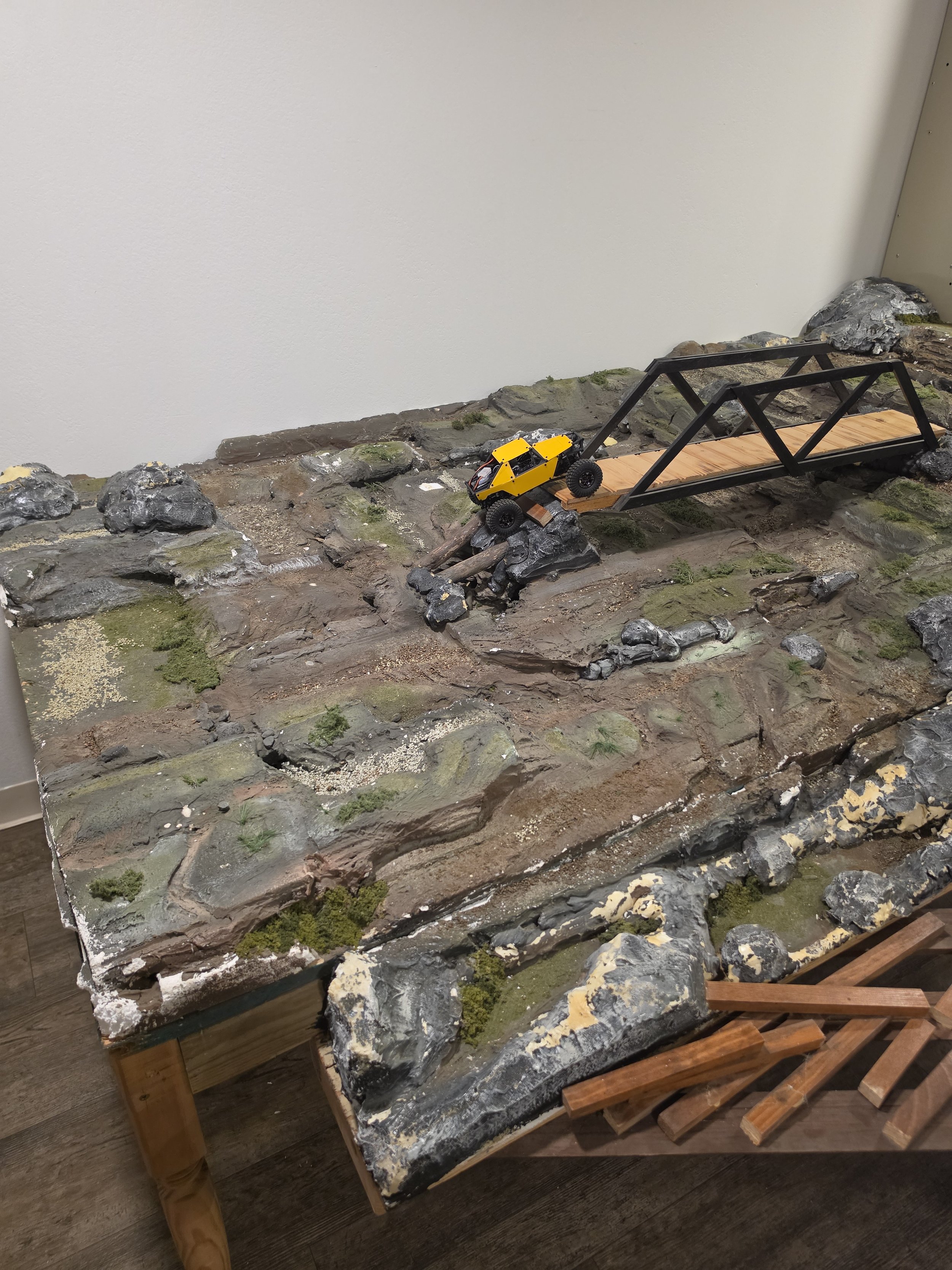 Small toy yellow and black off-road vehicle driving down a miniature rocky and earthy terrain with a small wooden bridge.