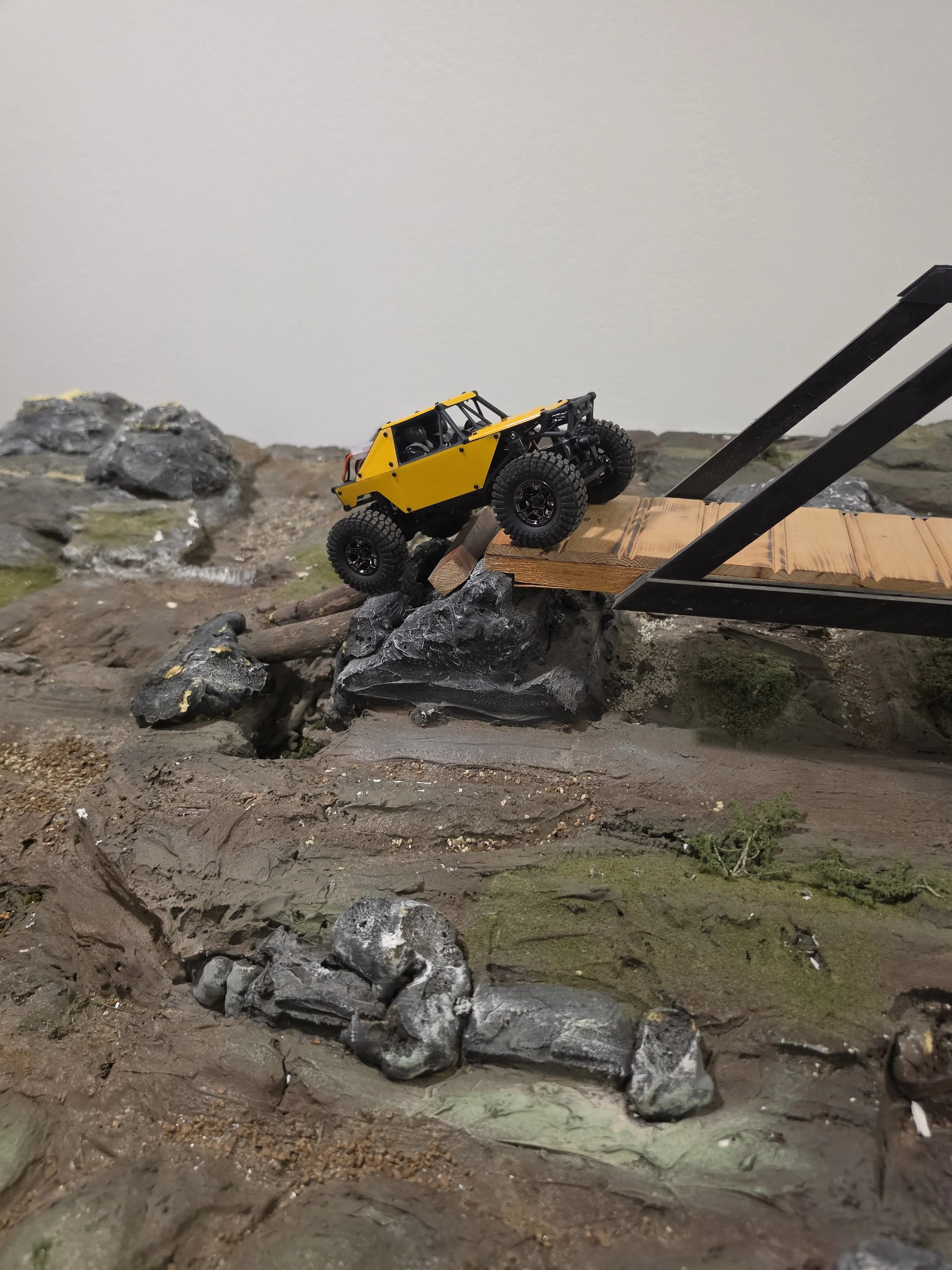 A small yellow and black off-road truck on a rocky terrain with a wooden bridge and black rocks.