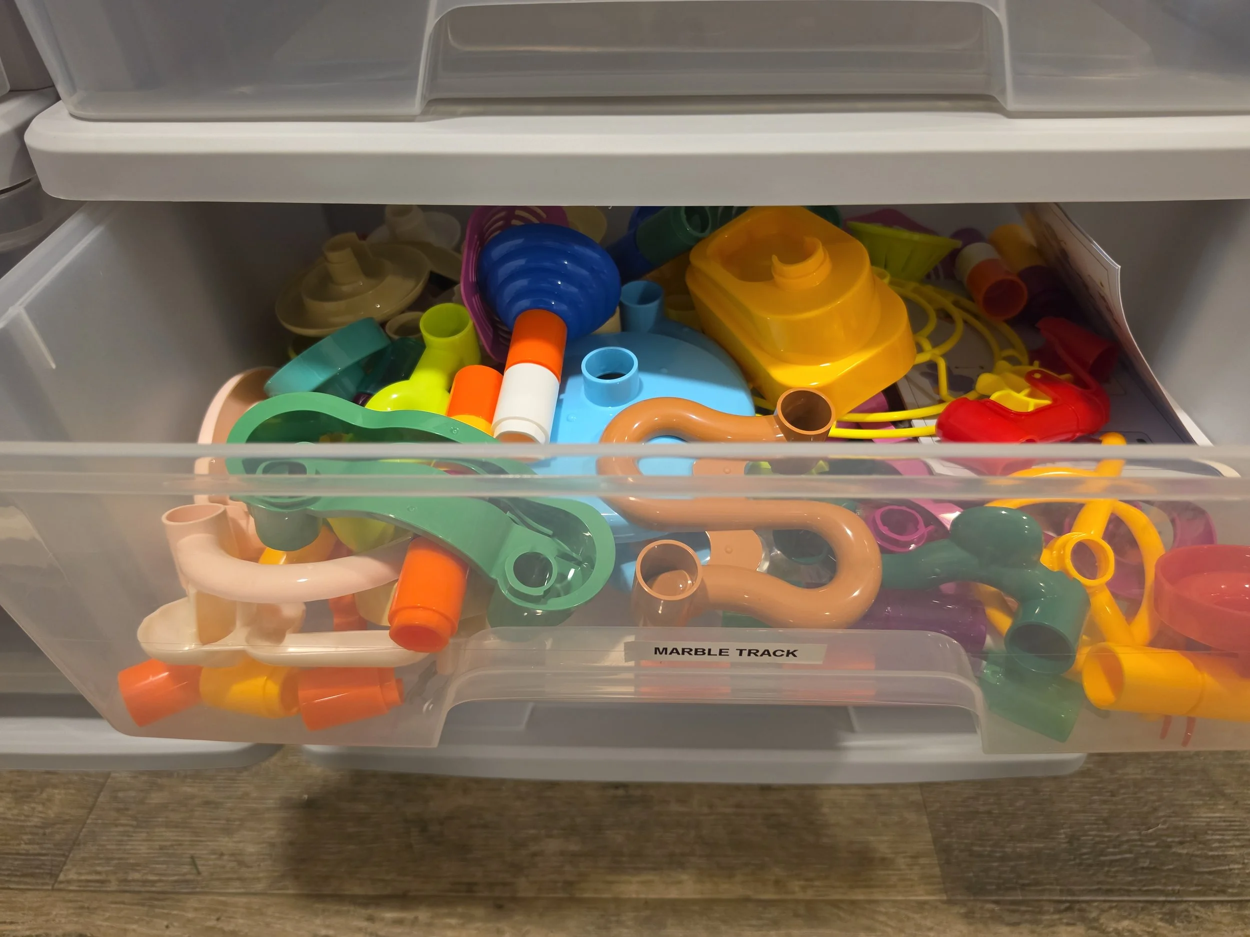 A clear plastic drawer labeled "Marble Track" contains colorful plastic toy parts, including tubes, connectors, and track pieces, in various bright colors like orange, yellow, green, blue, and pink.