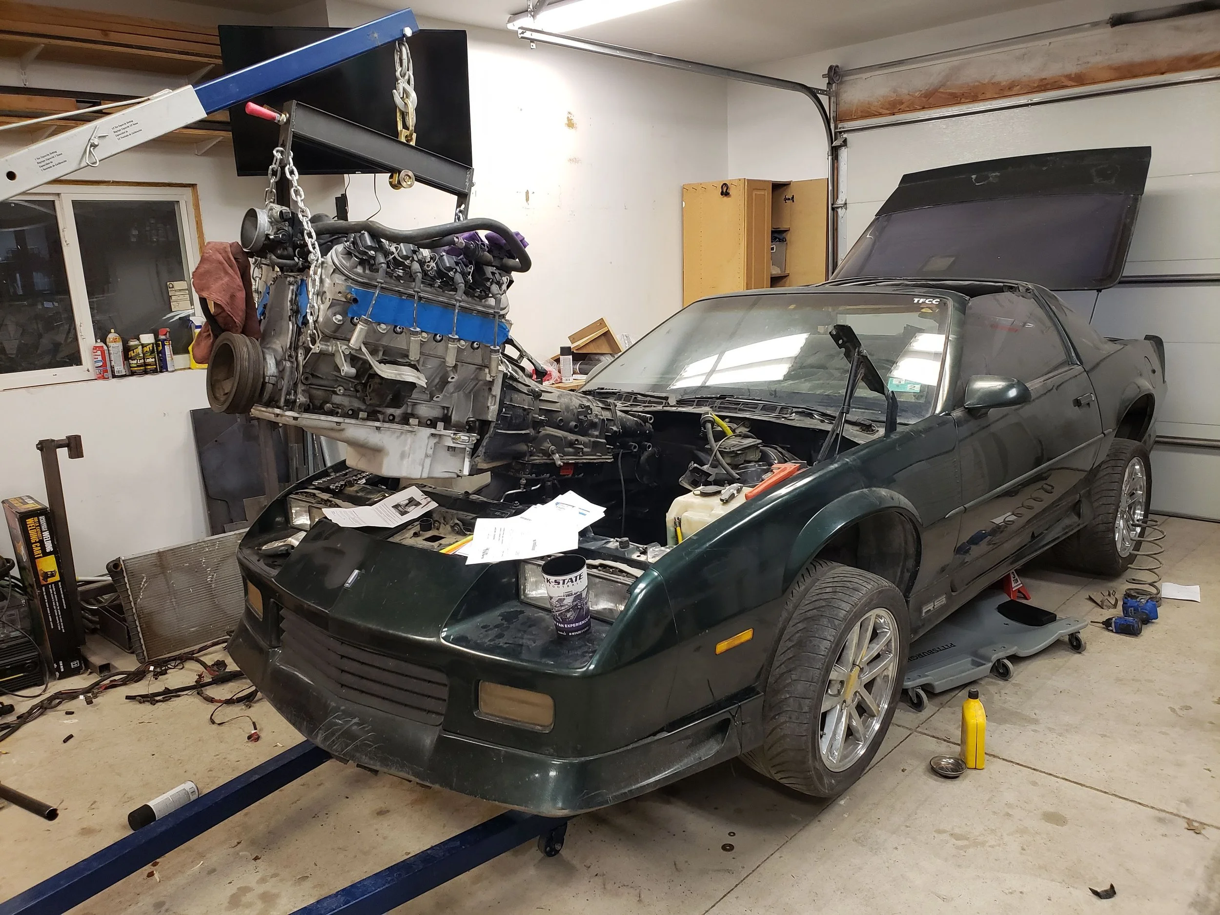 A garage with a car that is being repaired, with the front hood and engine removed. The engine is suspended in the air by a hoist. The garage has tools, parts, and supplies scattered around.