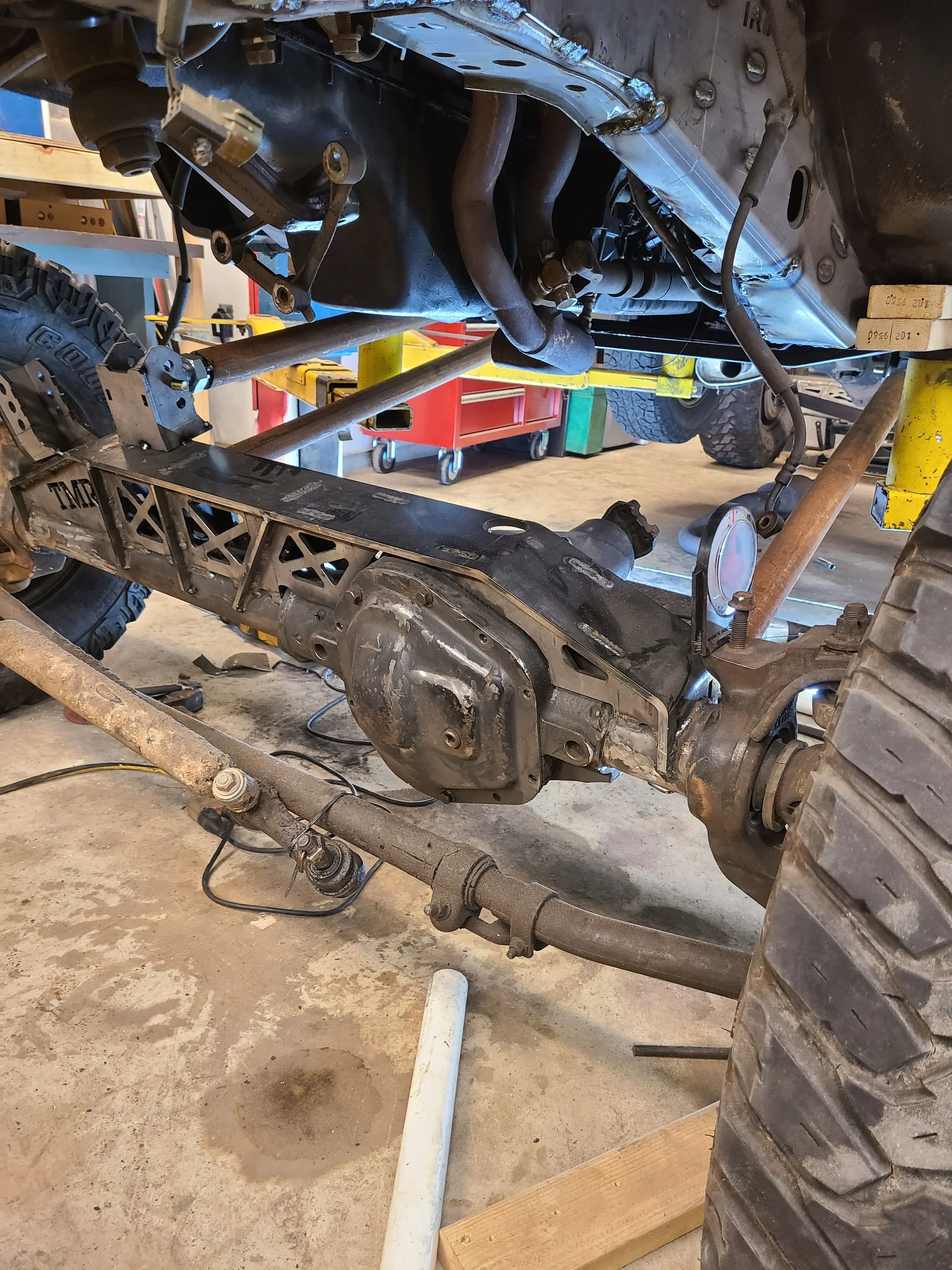 Underside of a vehicle in a garage or workshop, showing part of the chassis, suspension, drive shaft, and differential, with tools and workbenches in the background.