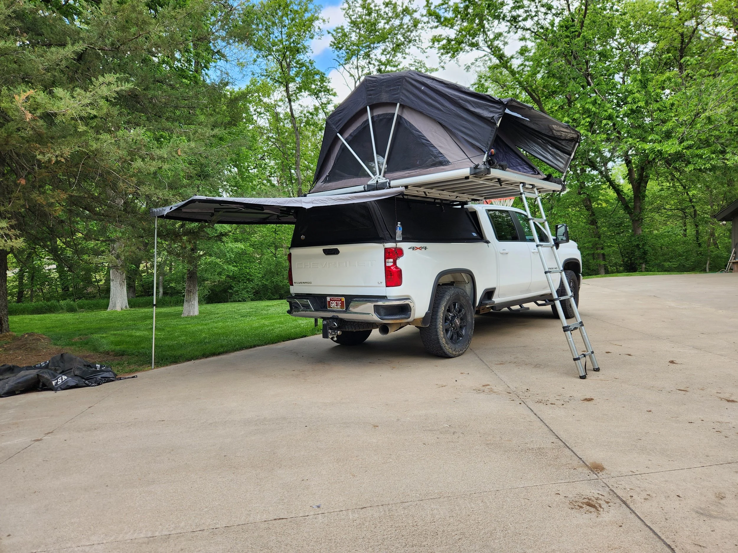 A white Chevrolet Silverado pickup truck with a roof-mounted tent and a slide-out extension parked on a paved driveway amidst green trees and grass.