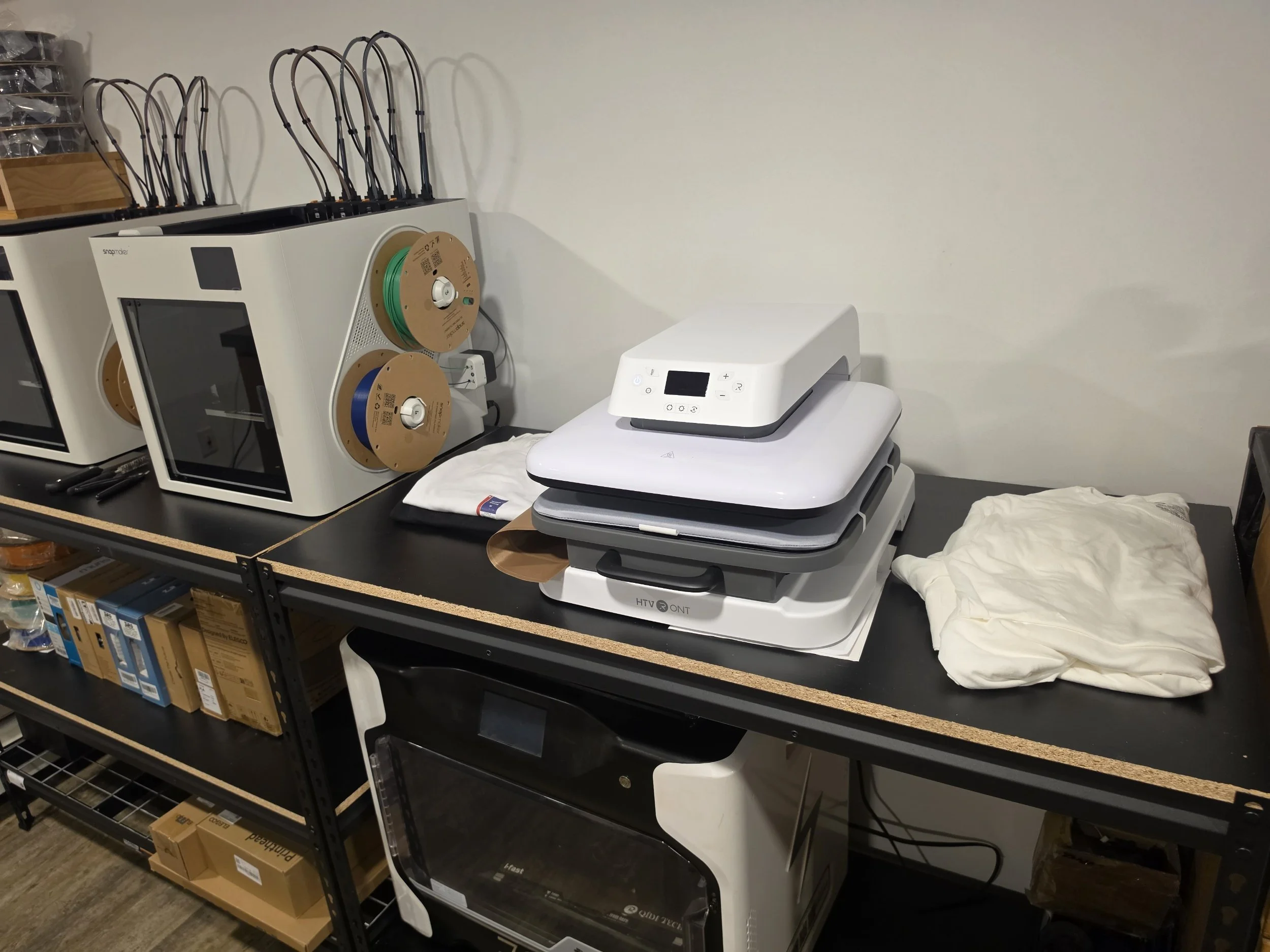A workbench with 3D printers, spool of filament, and a t-shirt press.