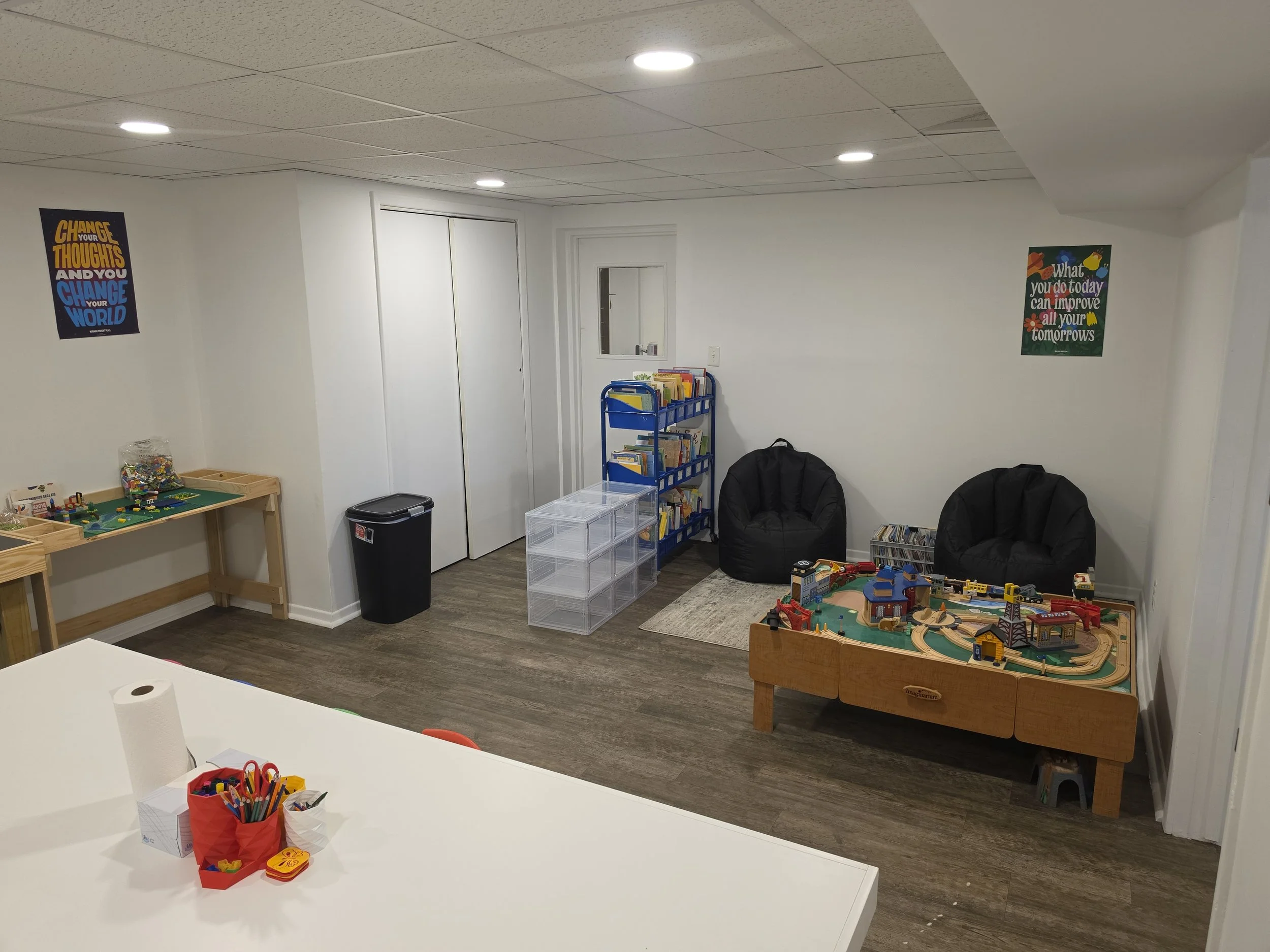 A playroom with white walls and ceiling tiles, decorated with motivational posters. There are two black bean bag chairs, a blue bookshelf filled with books, and various toys, including a train set on a wooden table. A small white desk with craft supp