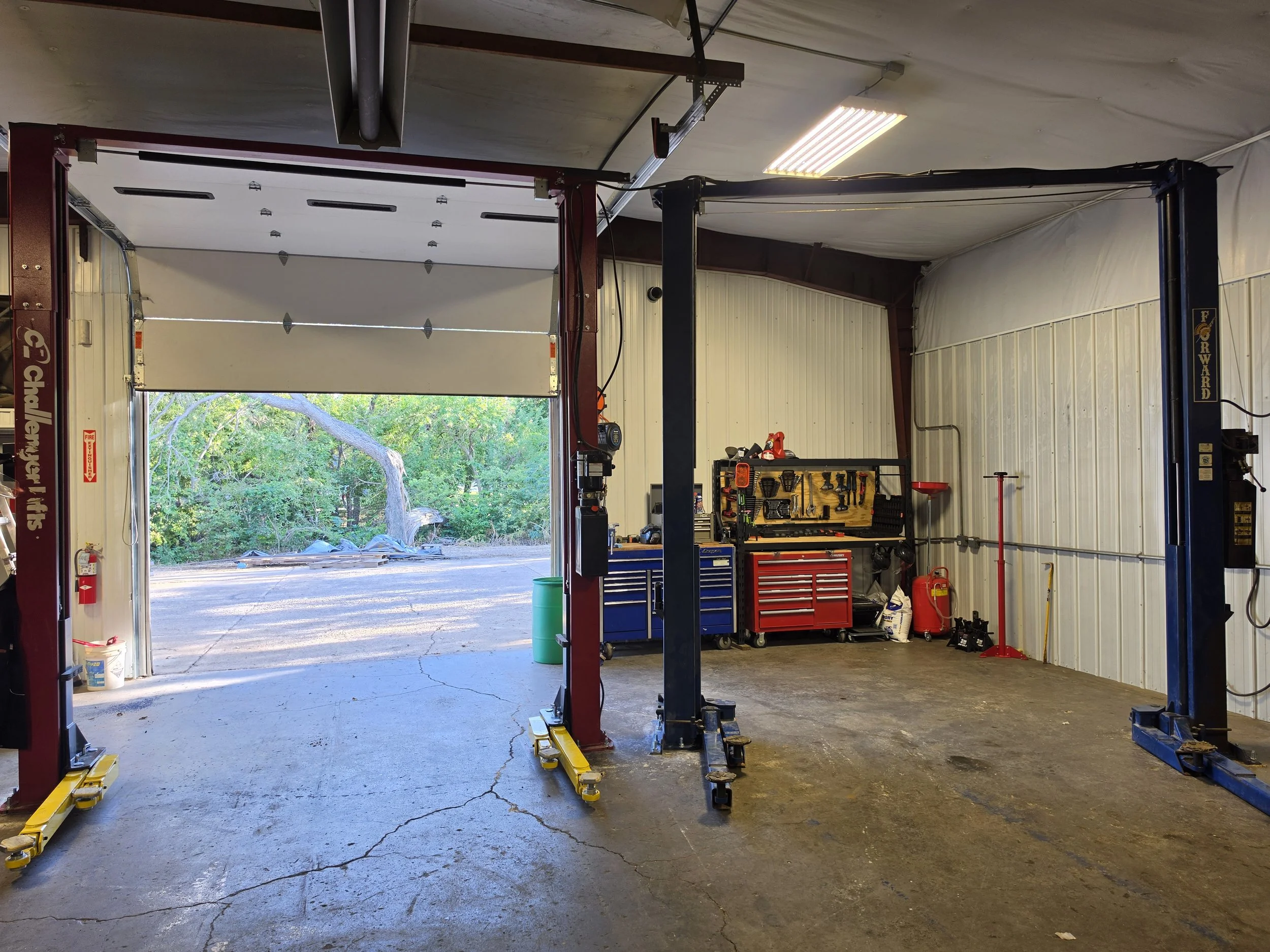 Inside a garage with an open garage door leading outside, showing trees and foliage. The garage contains a lift, toolboxes, and various tools and equipment.
