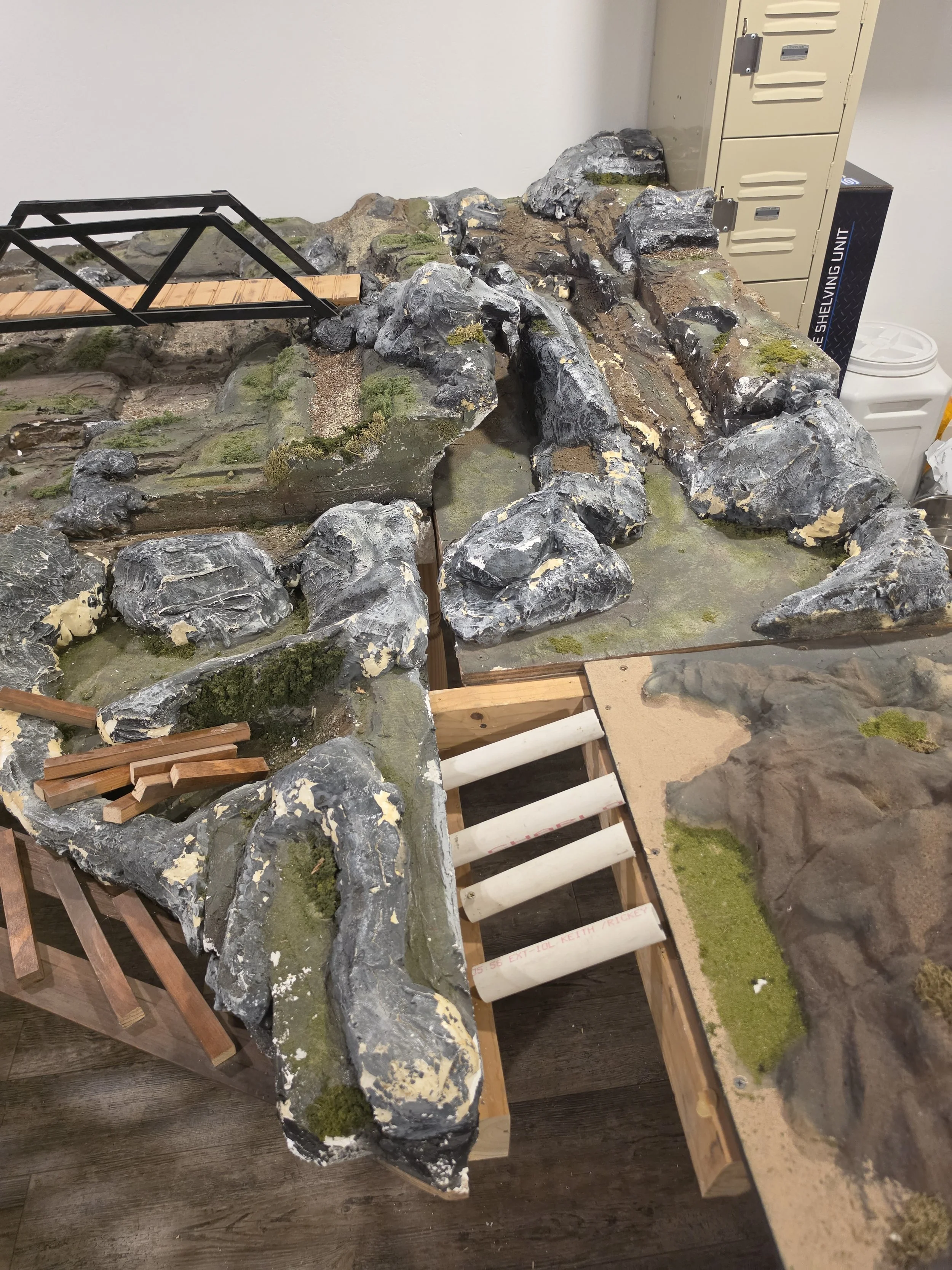 A detailed model landscape of rocks, terrain, and a small wooden bridge in an indoor setting, possibly a model train or diorama setup, with storage lockers and other objects in the background.