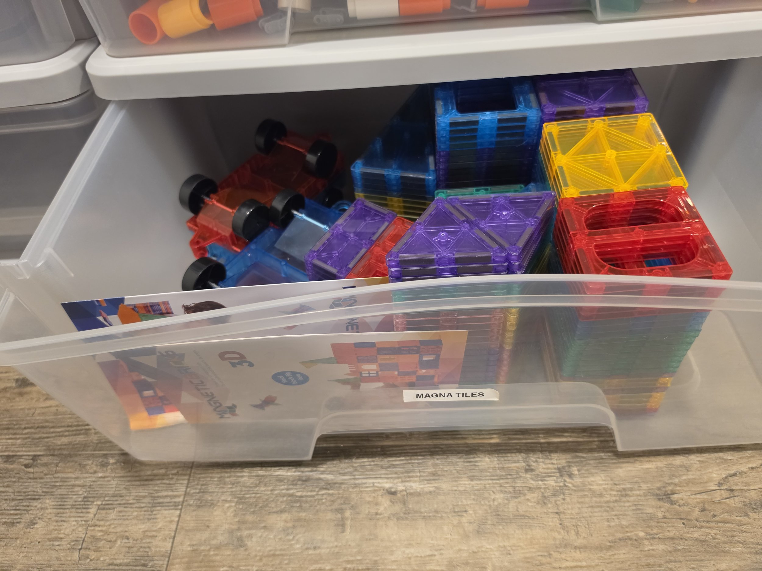 A clear plastic storage bin labeled 'MAGNA TILES' contains colorful magnetic building tiles in red, yellow, purple, green, and blue, with some tiles stacked neatly.