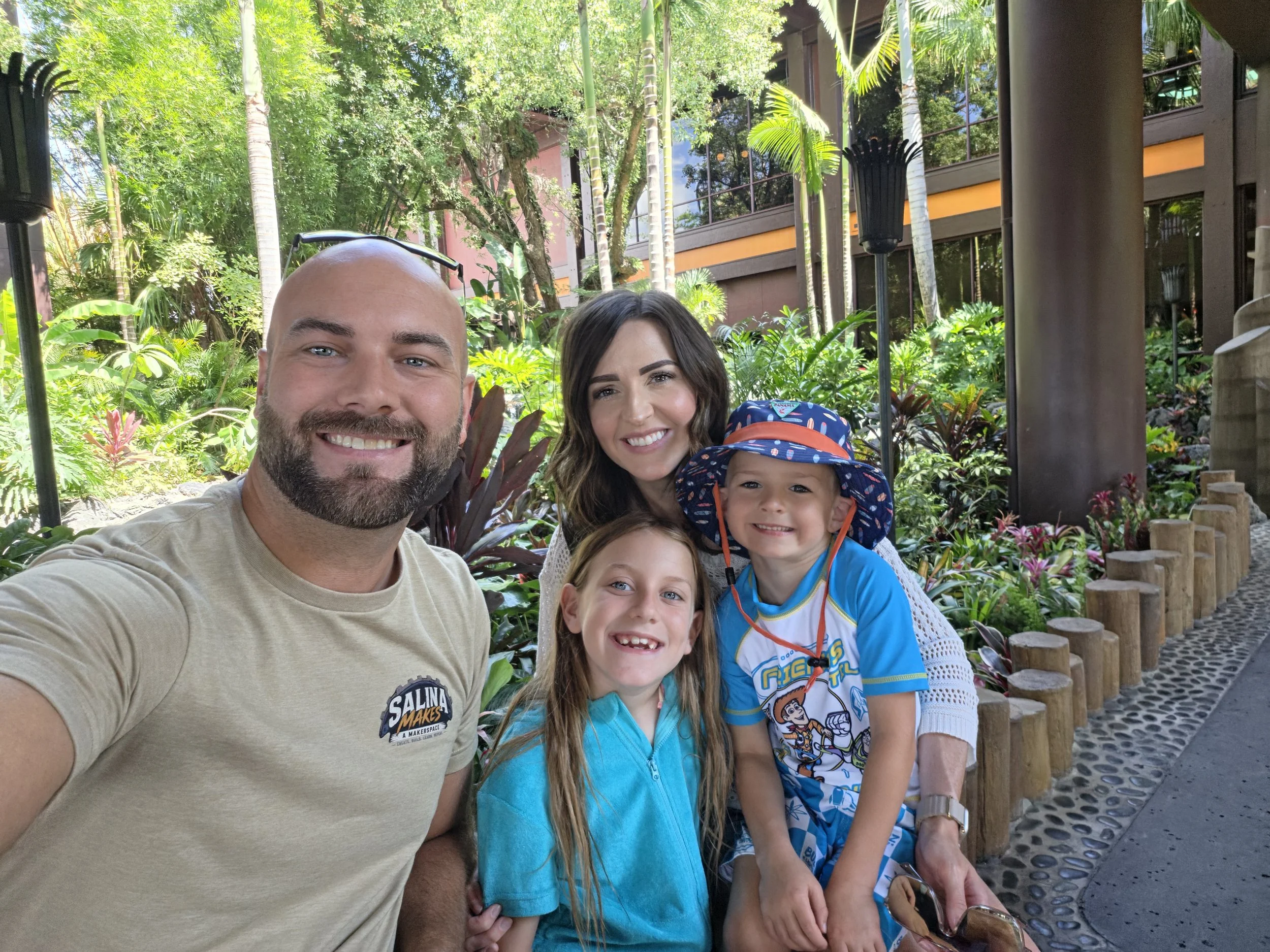 A smiling family of four taking a selfie outdoors amidst lush green tropical plants and trees, with modern buildings in the background.