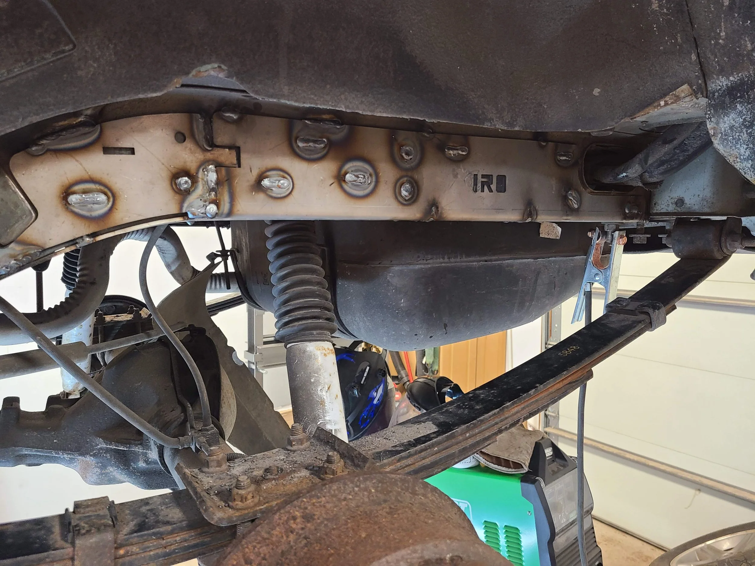 Close-up of a metal vehicle chassis showing weld marks, rust, and parts of suspension and wiring.