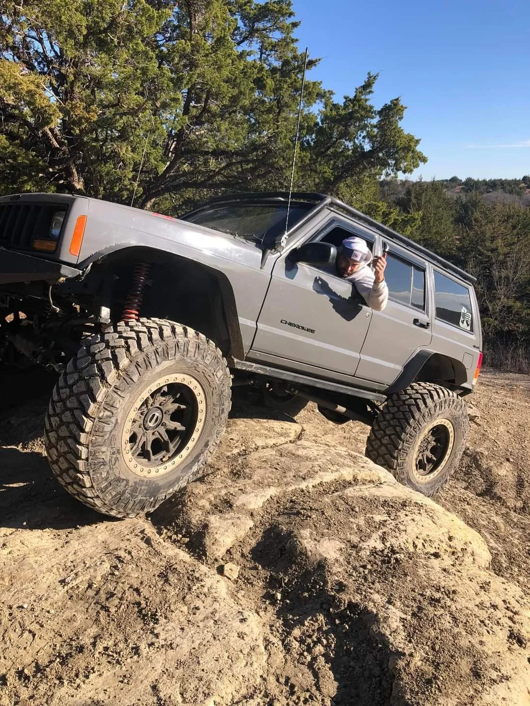 A person inside a gray Jeep Cherokee off-road vehicle navigating rocky terrain, with large rugged tires and a tree in the background.