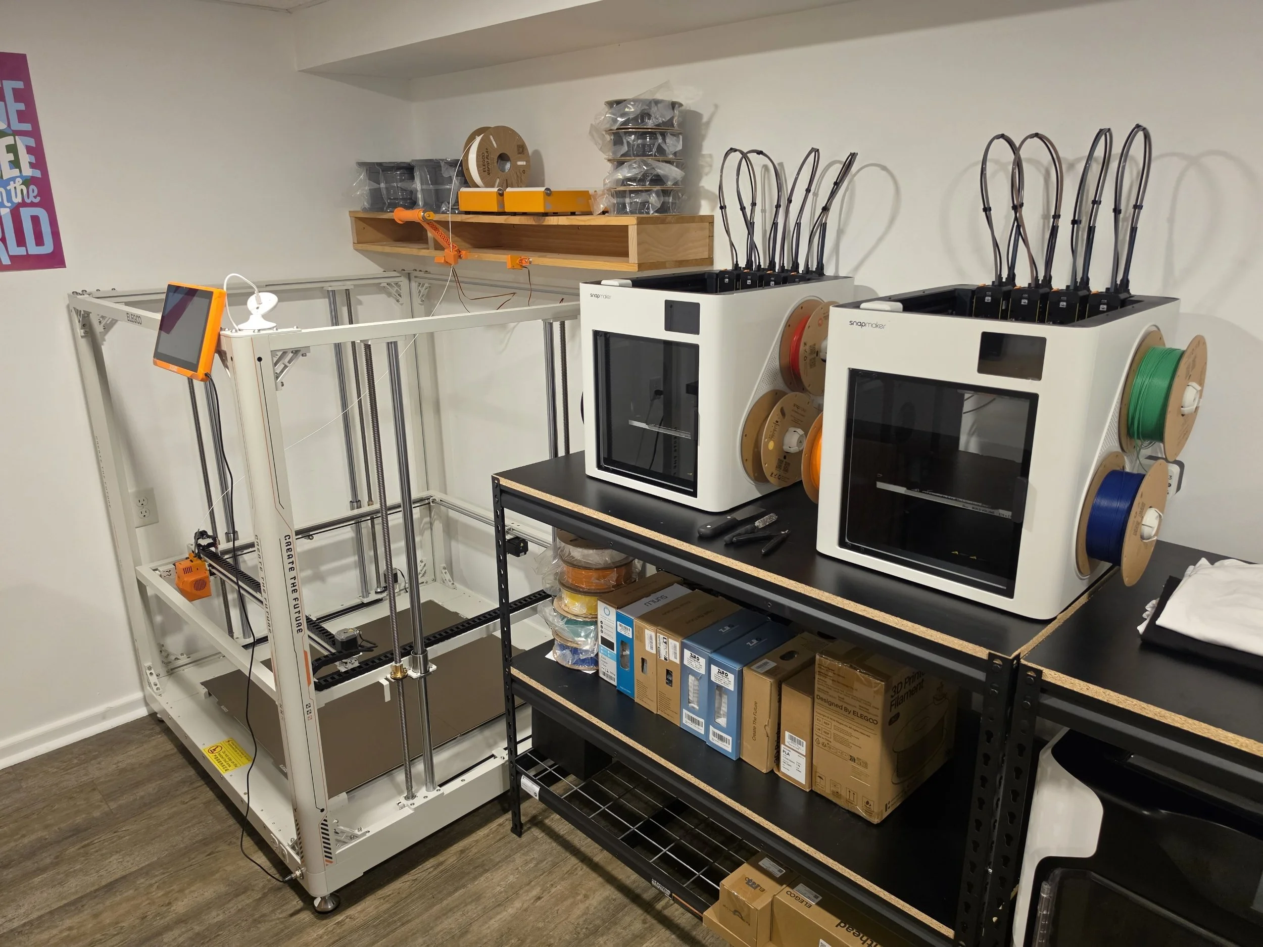 A room with electronics and 3D printing equipment. On the right, there are spools of filament in various colors on a black shelving unit. In the middle, there are two white 3D printers with multiple black wires connected to them. To the left, an empty white 3D printer frame is visible, and some tools are placed on a black workbench. The background features a beige wall with a pink and blue poster partially visible on the left.