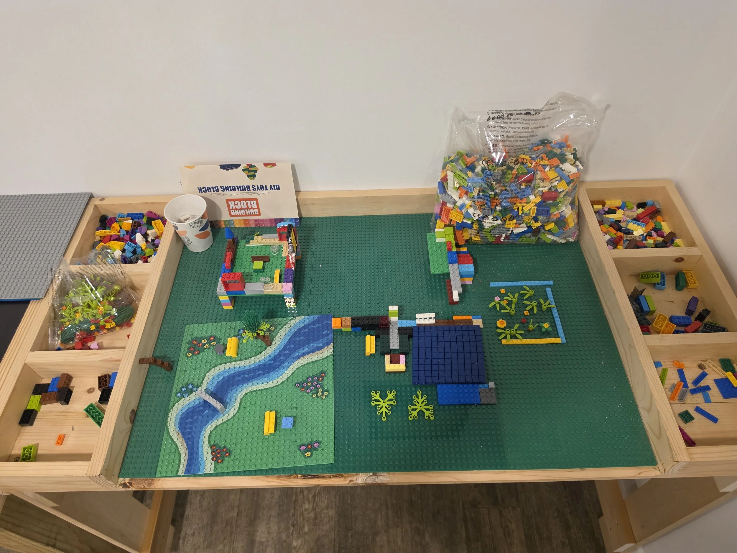 A LEGO construction table with a partially built landscape using green baseplates, featuring a river made of blue LEGO bricks, small trees, and flowers. Surrounding the table are containers with colorful LEGO bricks, a box labeled 'DIY YOUR BUILDING 