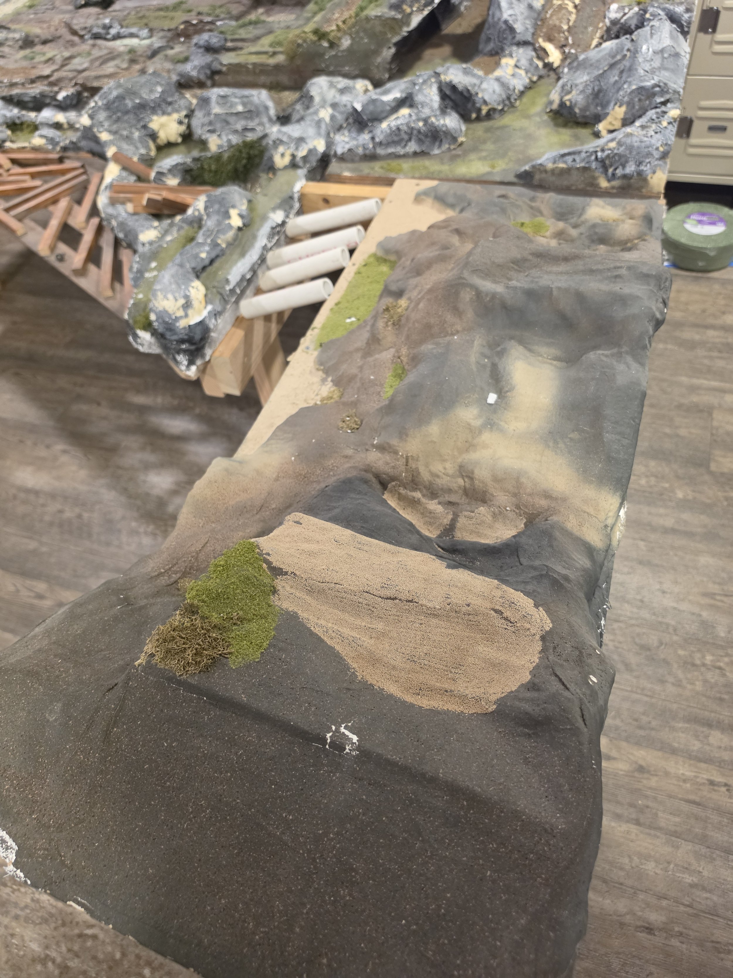 A close-up view of a miniature landscape with rocks, moss, and sand on a wooden surface, likely part of a model or diorama.