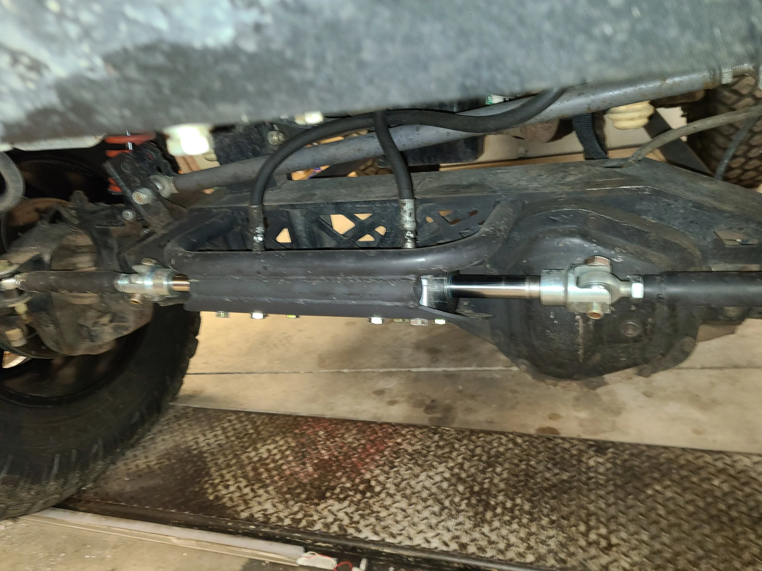A close-up view of a motorcycle's lower control area showing parts of the frame, suspension, and tires. The image is taken from underneath the motorcycle, highlighting the mechanical components.