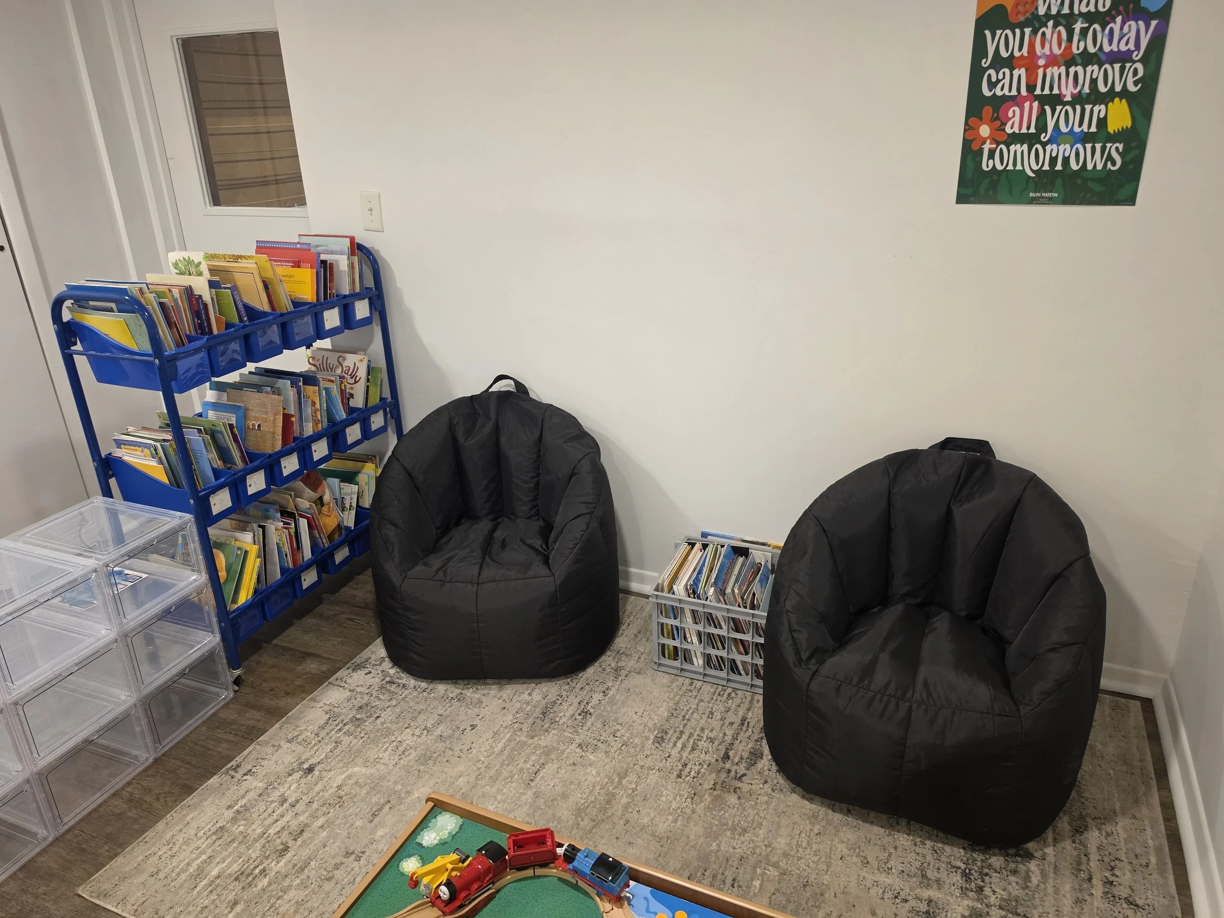 A room with a white wall decorated with a colorful inspirational quote poster that says, 'What you do today can improve all your tomorrows.' There is a blue bookshelf filled with books, a clear plastic storage container, and two black bean bag chairs