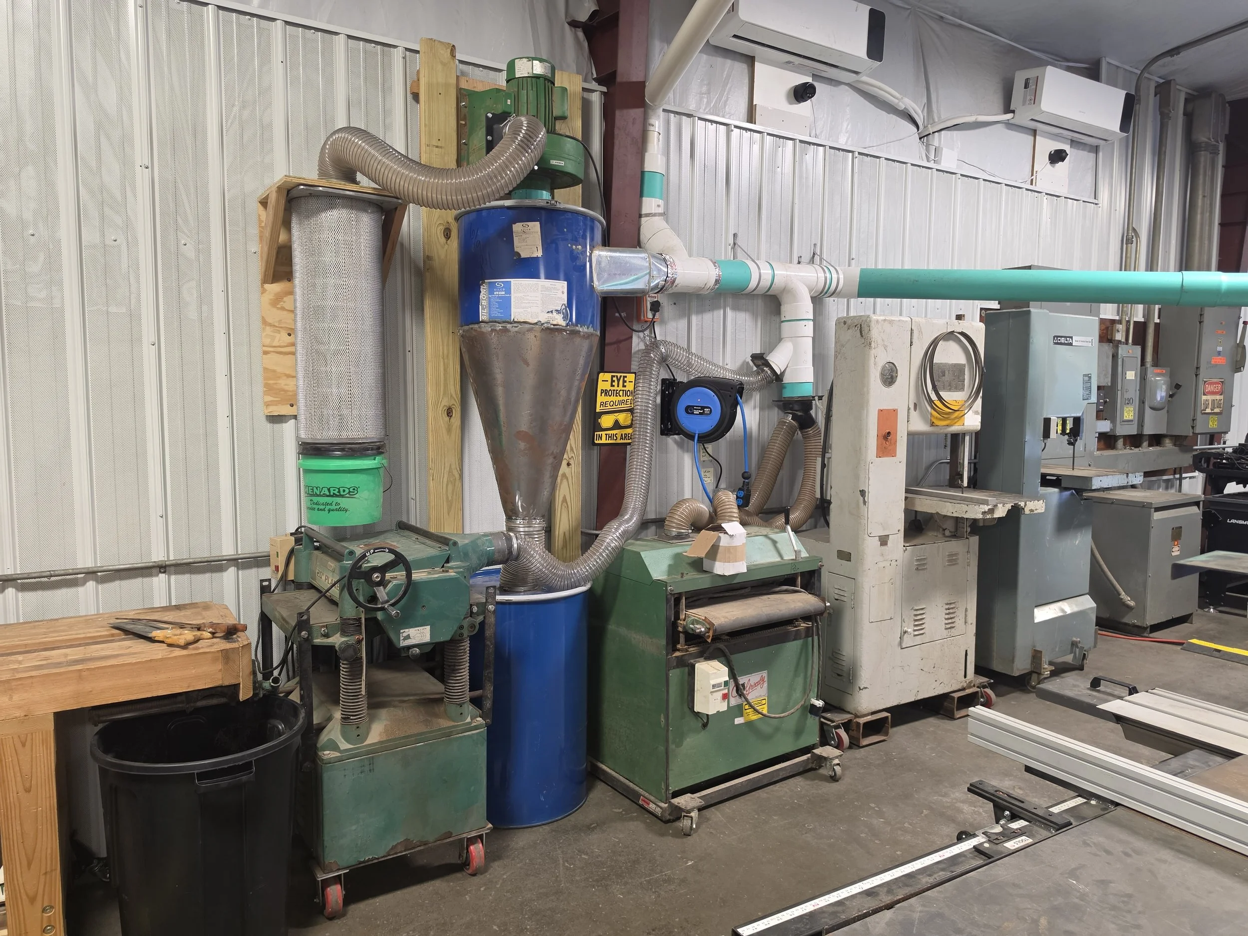 Workshop with various woodworking equipment and tools, including a dust collector, workbenches, and machines, set against a metal wall.