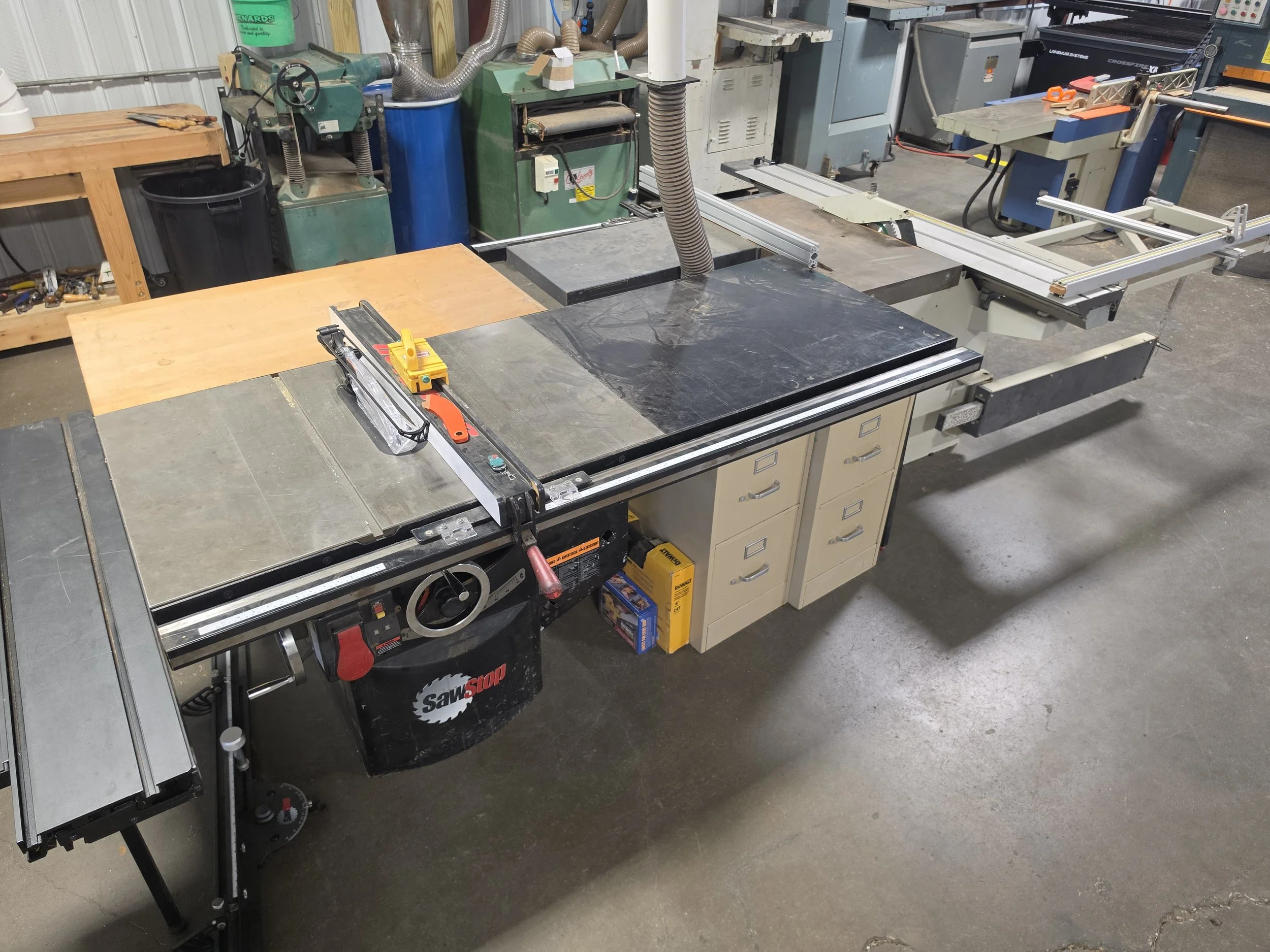 A workshop with various woodworking and machinery equipment, including a table saw, workbenches, and dust collection hoses.
