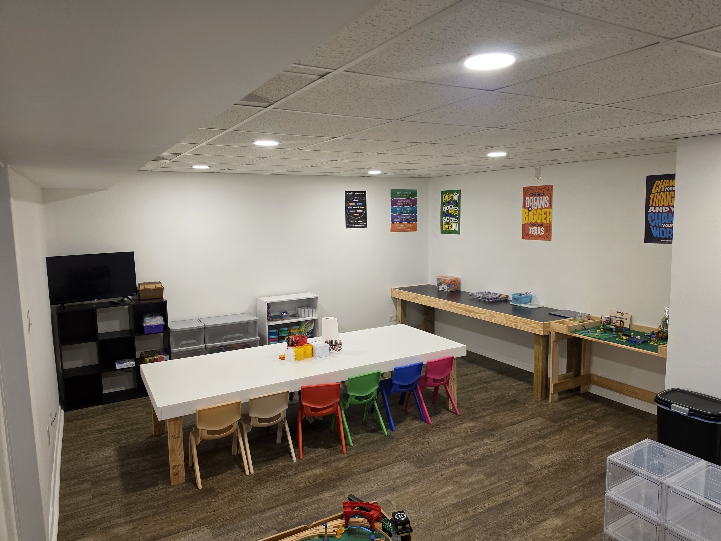 An indoor playroom or classroom with colorful chairs around a white table, a black shelving unit with storage bins, a small white cabinet, and posters with motivational messages on the walls. There are also wooden tables and clear plastic bins for to