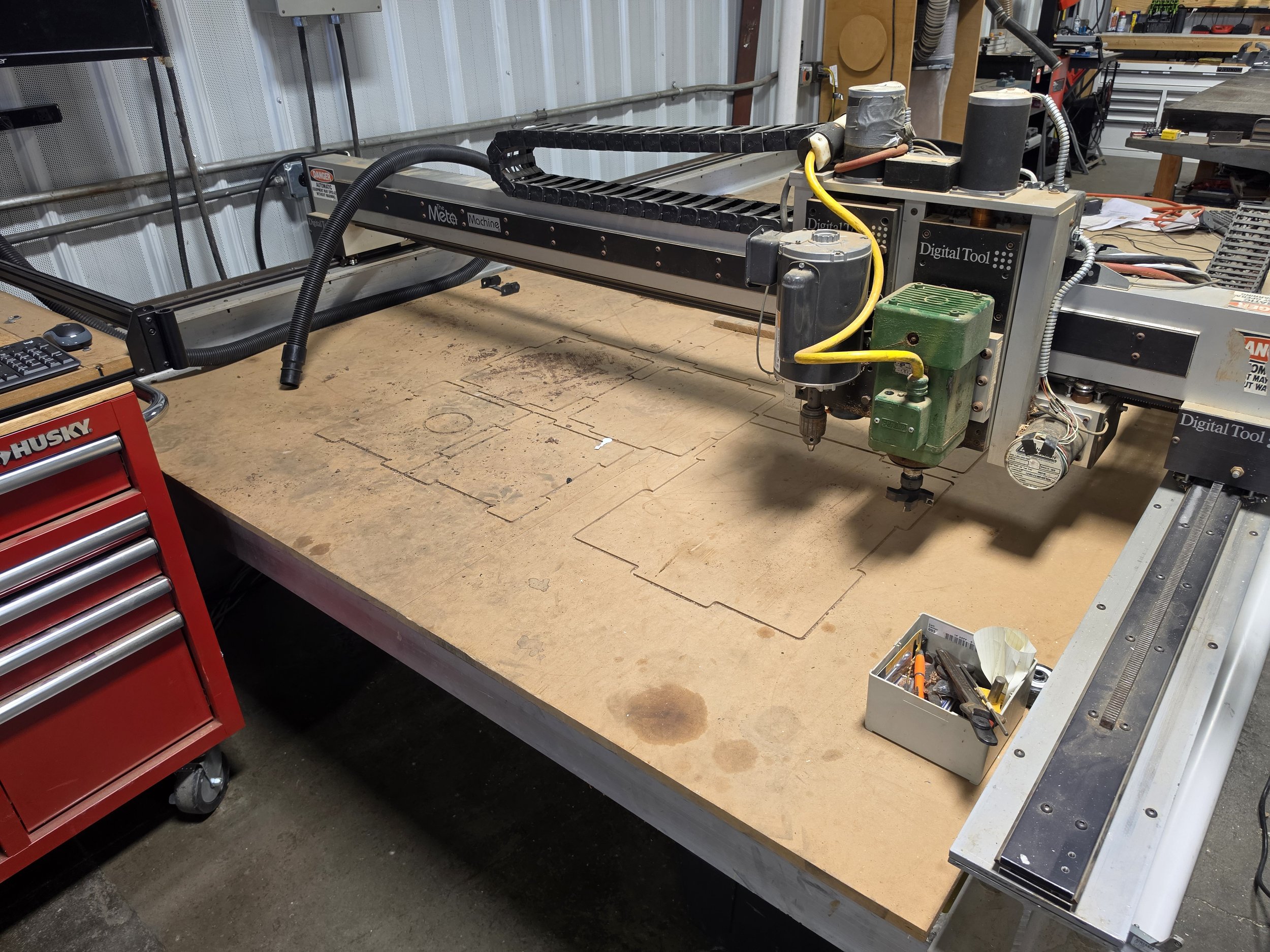 A workshop table with a large CNC machine, tools, and equipment. A red toolbox is placed at the left corner, and there are various tools and materials on the machine and nearby surfaces.