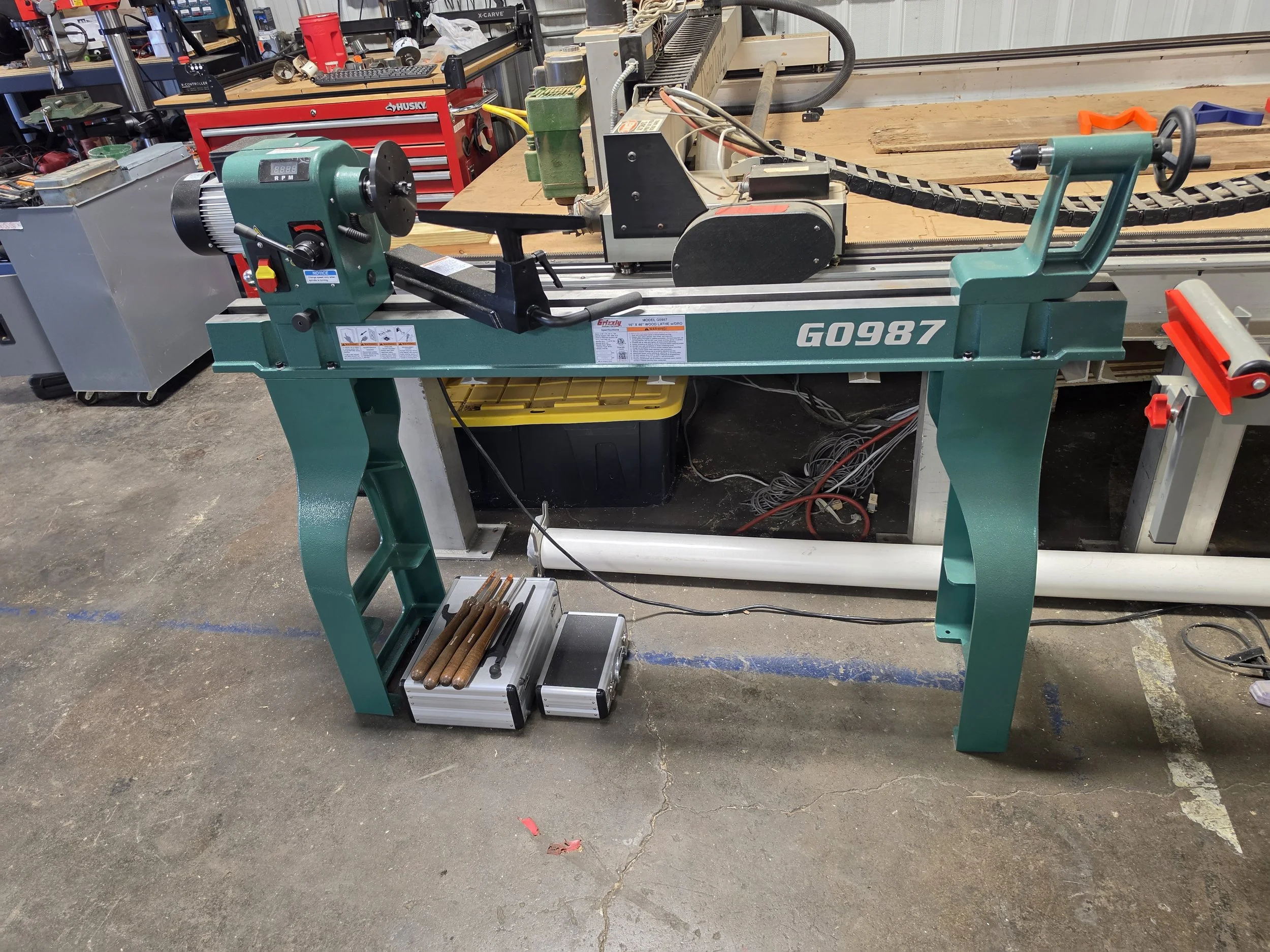 A woodworking lathe machine in a workshop. The lathe is turquoise with controls and a rotating spindle for shaping wood. There are various tools and equipment in the background, along with a workbench and storage drawers.
