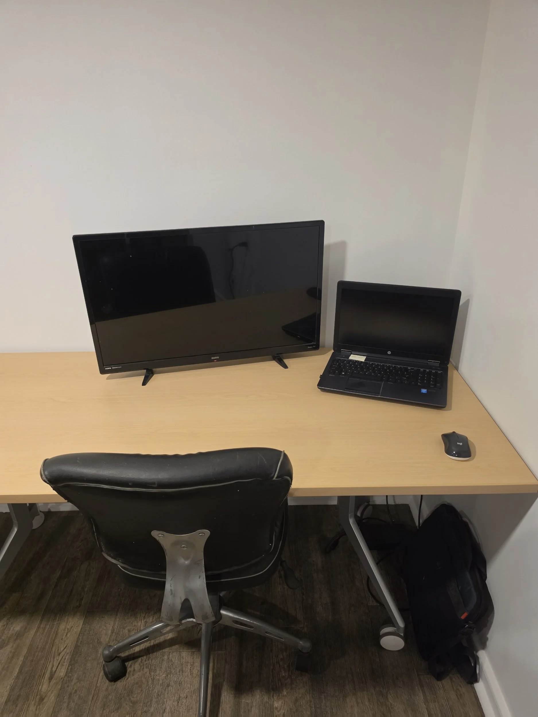 A simple office workspace with a wooden desk. On the desk, there is a large black monitor, a black laptop, and a small wireless mouse. A black office chair is in front of the desk. Under the desk, there is a black backpack. The background wall is plain and white, and the floor is made of dark wooden planks.
