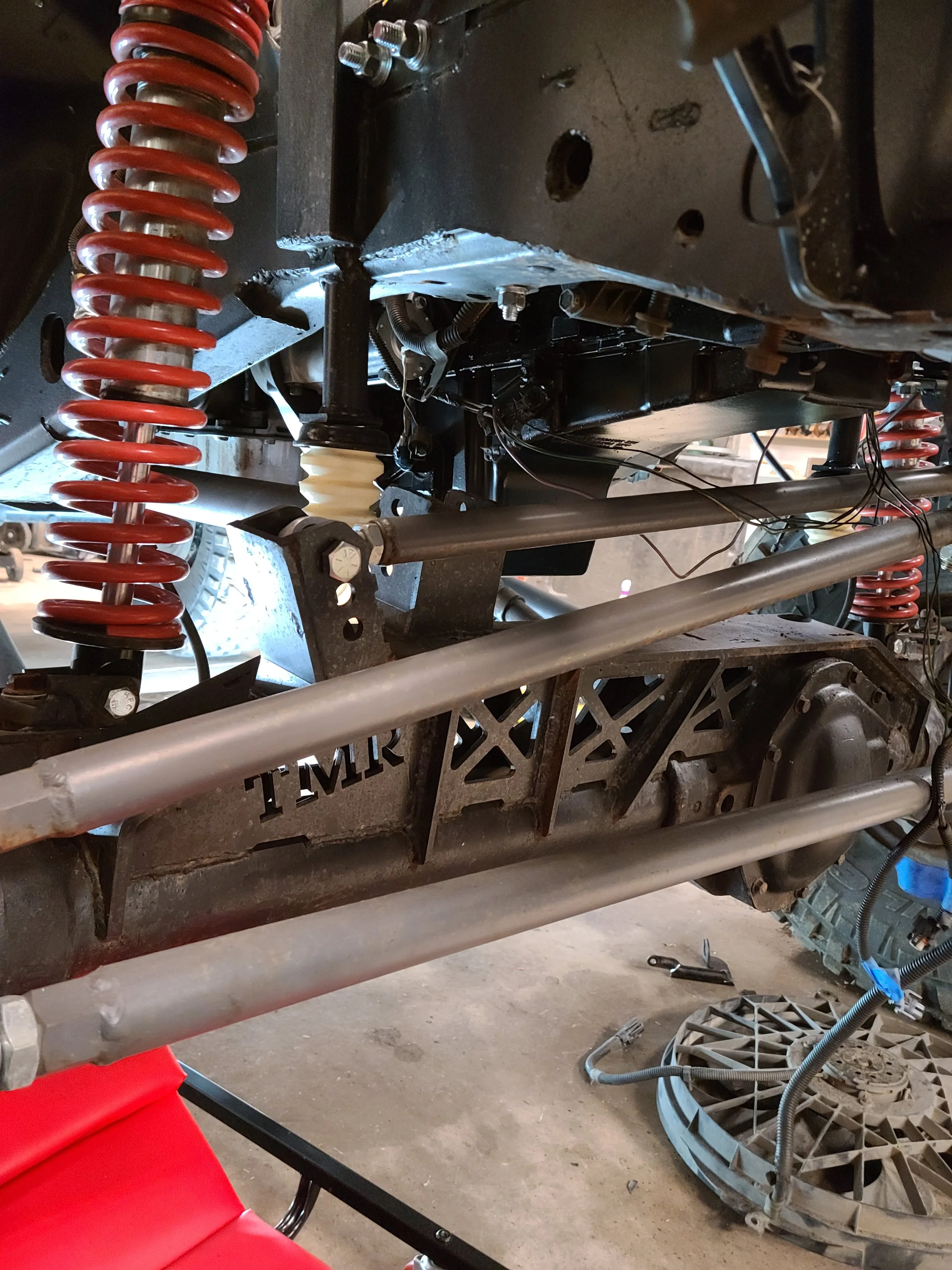 Close-up of a vehicle chassis with suspension components, including a red coil spring, a black shock absorber, and various metal rods and bolts, in a workshop setting.