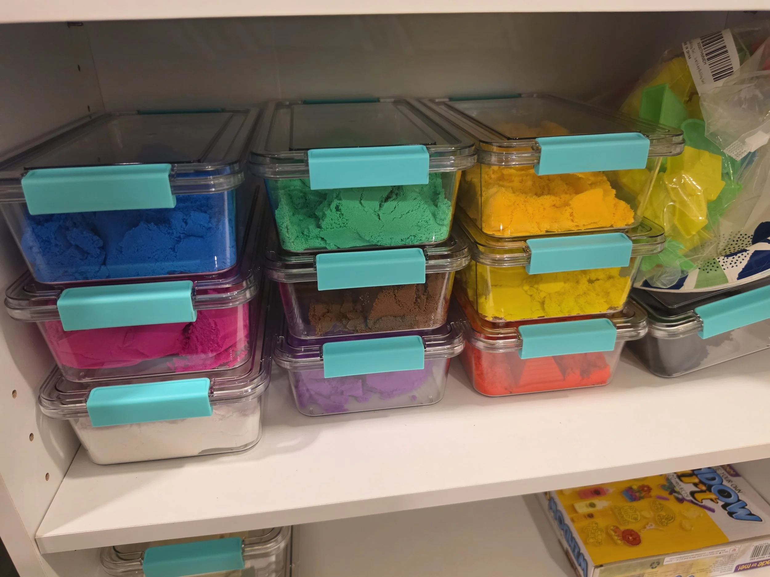 Shelf with multiple plastic containers filled with colored powders, including blue, green, yellow, purple, brown, and white, used for arts and crafts.