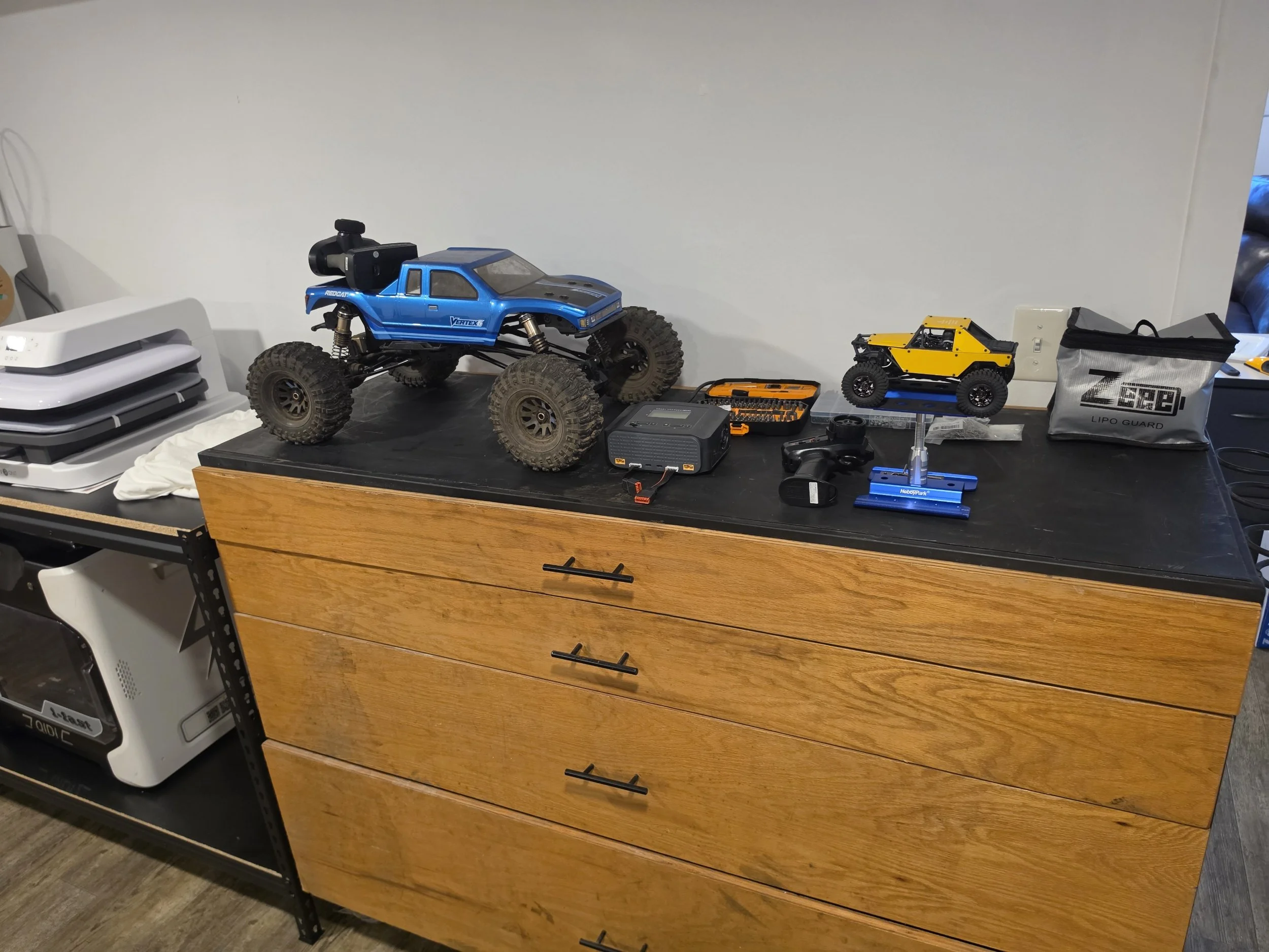 A desk with a blue remote-controlled off-road vehicle, a smaller yellow remote-controlled vehicle, various tools, a charging station, and pouches on a wooden drawer unit, with stacked electronic devices on a separate stand.