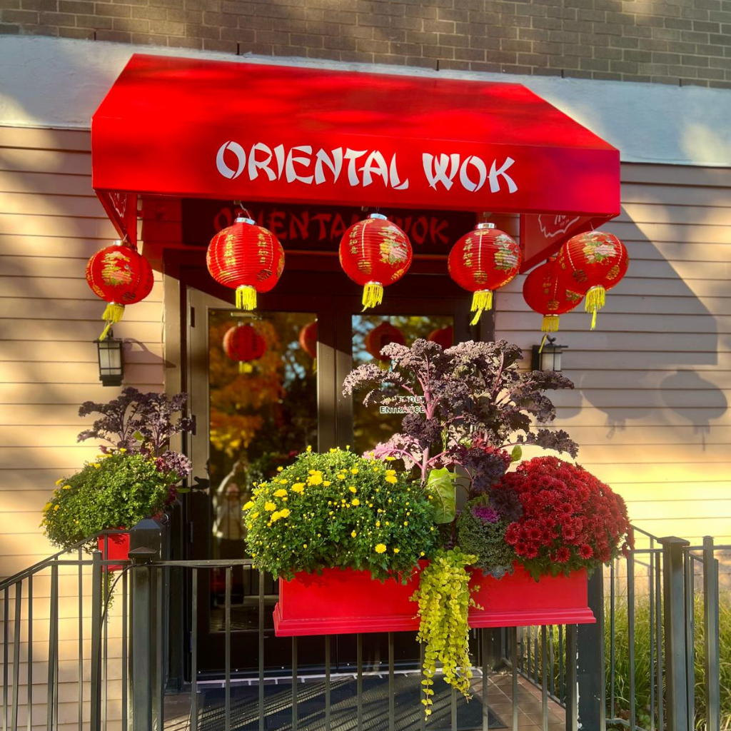 Entrance of a restaurant named 'Oriental Wok' with a red awning, red lanterns, and colorful potted plants including yellow, purple, and red flowers outside.
