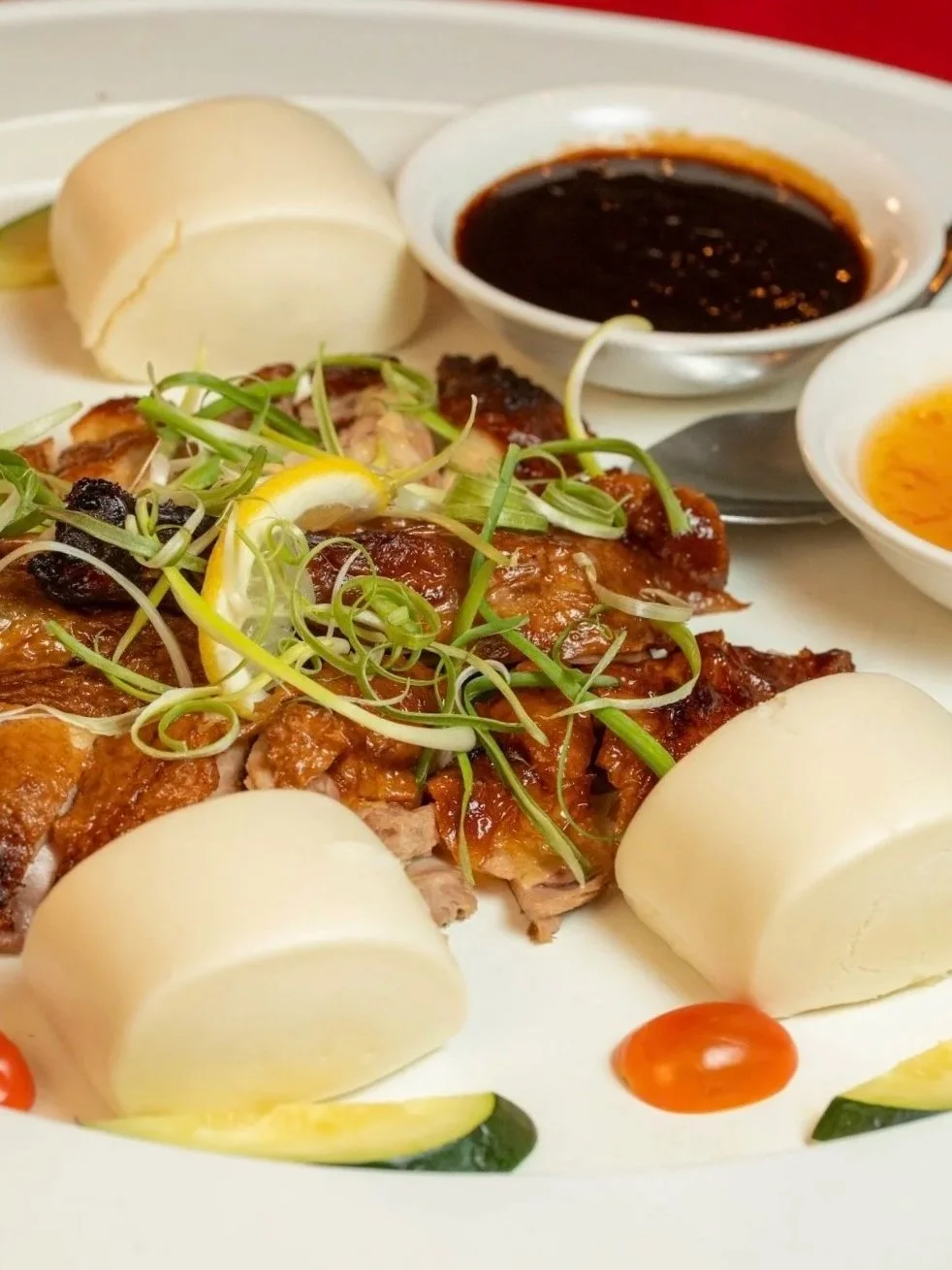 A plate of sliced boiled chicken with green onions and lemon slices, accompanied by steamed bun rolls and dipping sauces in small bowls.